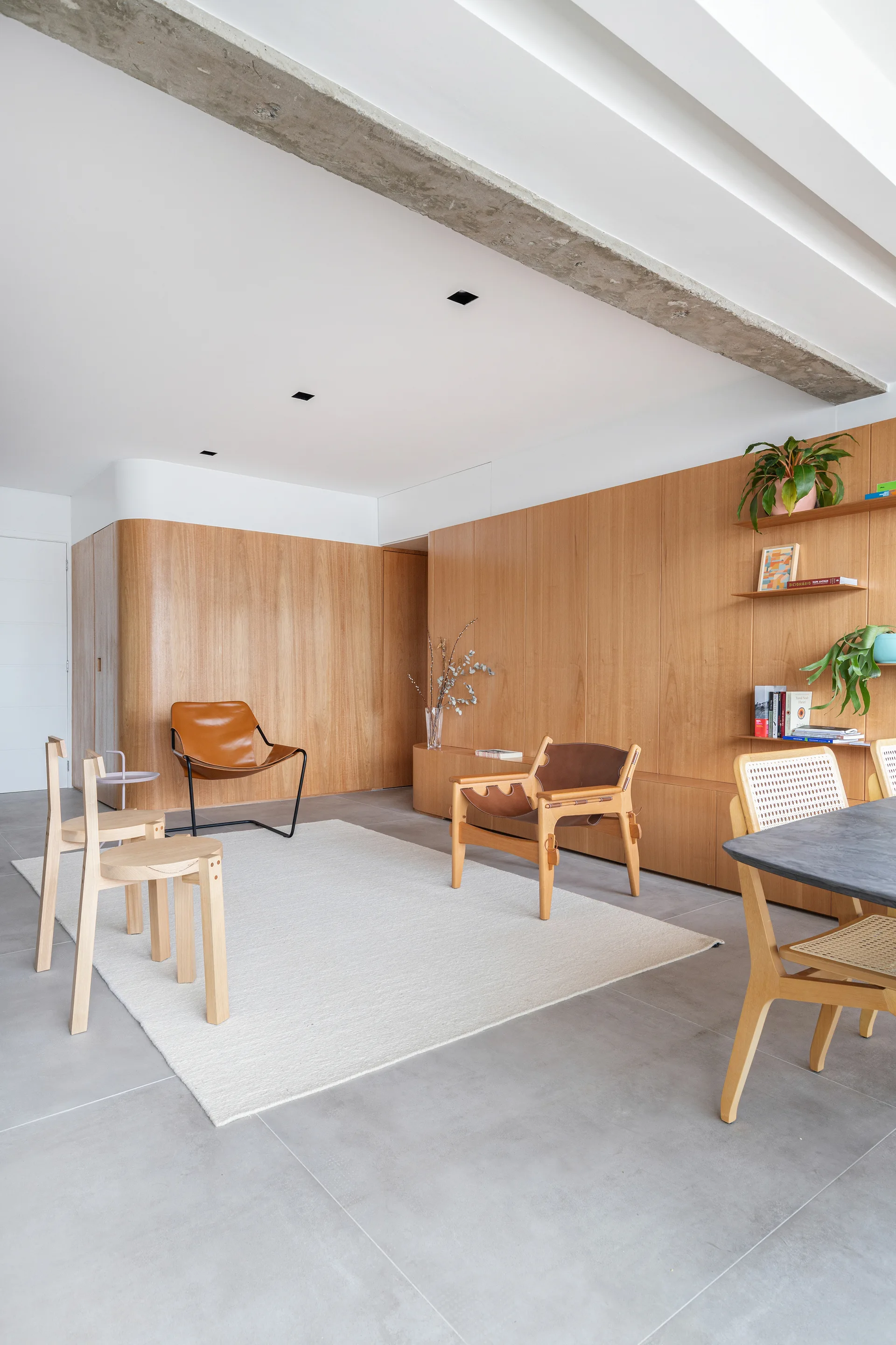 A modern living area features light wood wall paneling, minimalist leather armchairs on a white rug, and an exposed concrete ceiling beam.