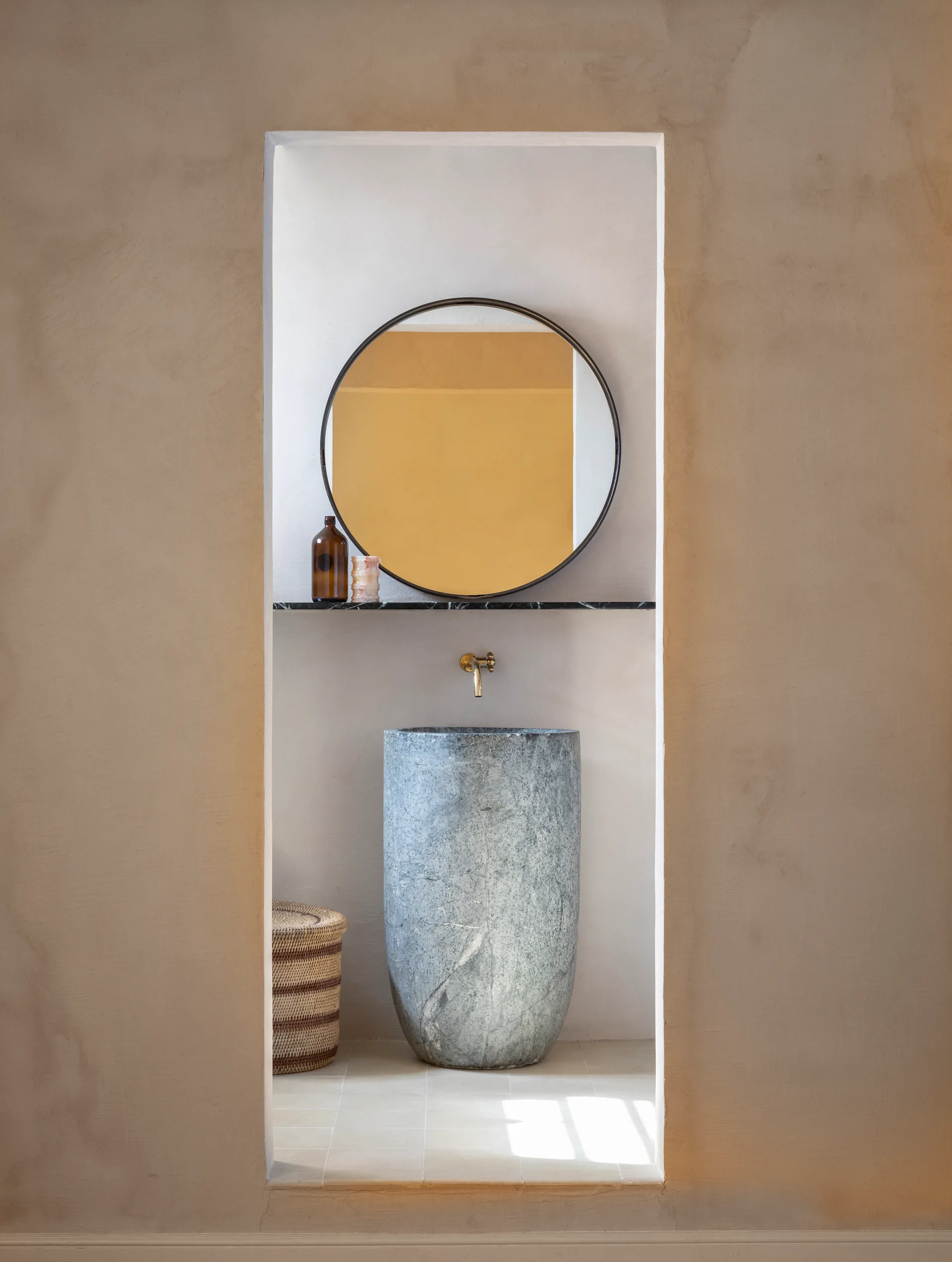 A minimalist bathroom features a textured plaster wall niche housing a tall grey stone pedestal sink, a brass faucet, and a circular mirror.