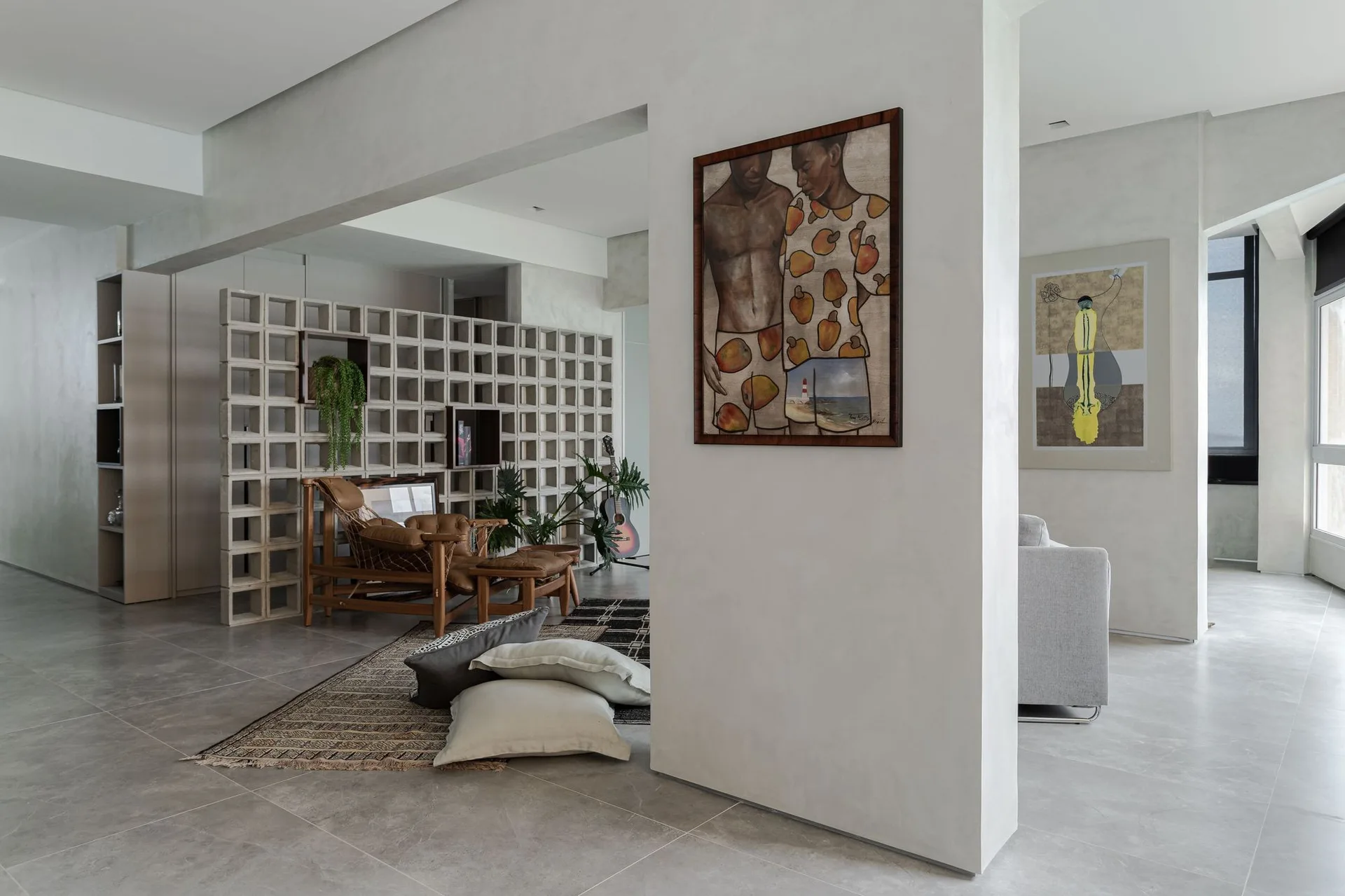 A living room features a concrete breeze block partition, a brown leather lounge chair on layered rugs, and a structural column with a painting.