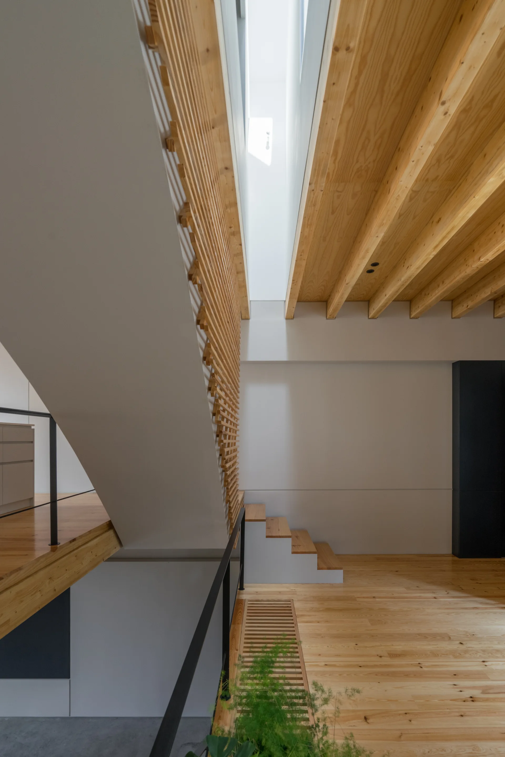 A modern wooden staircase features a vertical slatted timber screen, exposed ceiling beams, and bright natural light from an overhead skylight.