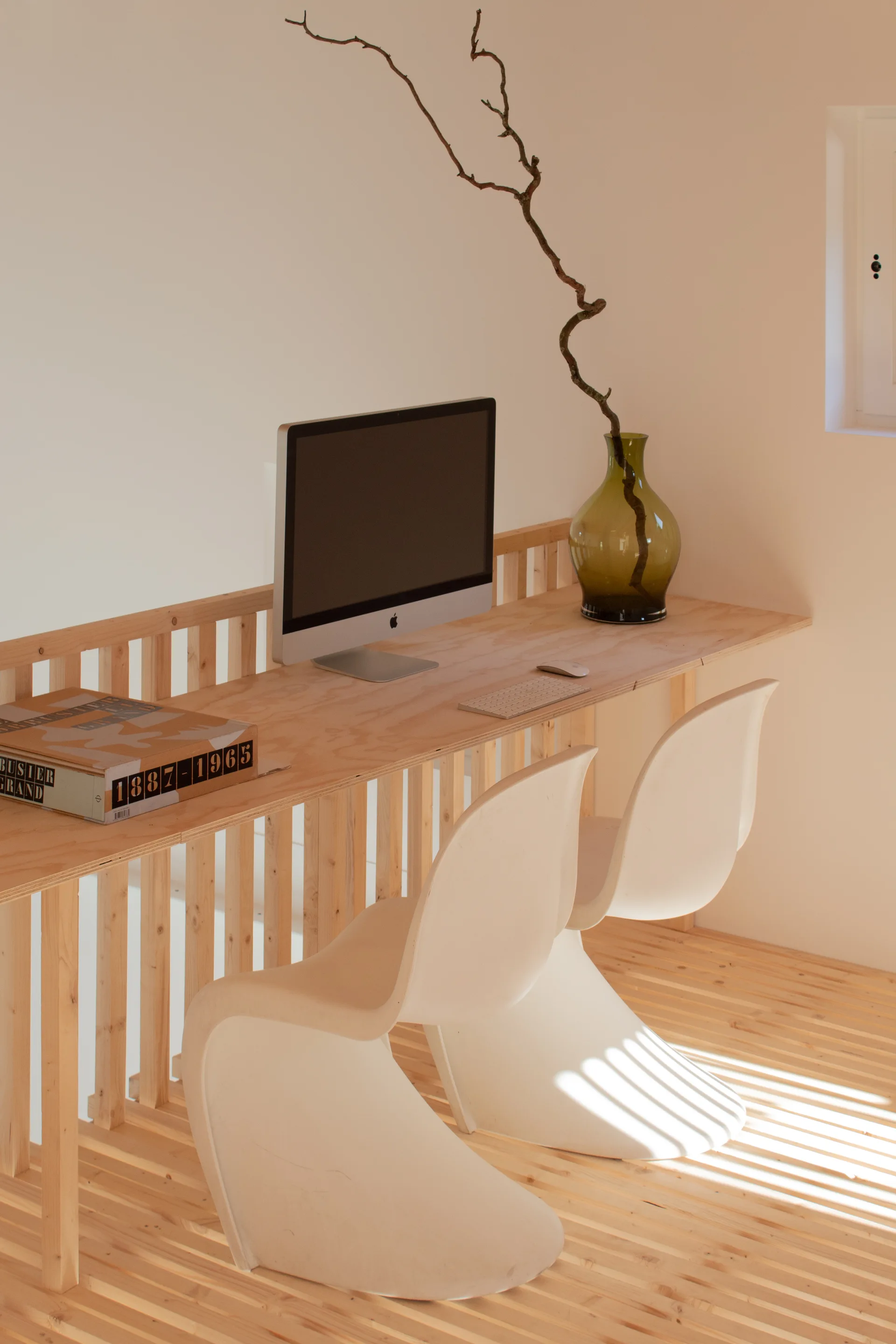 A minimalist plywood desk built into a wooden slat railing features a desktop computer, a green glass vase, and two white sculptural chairs.