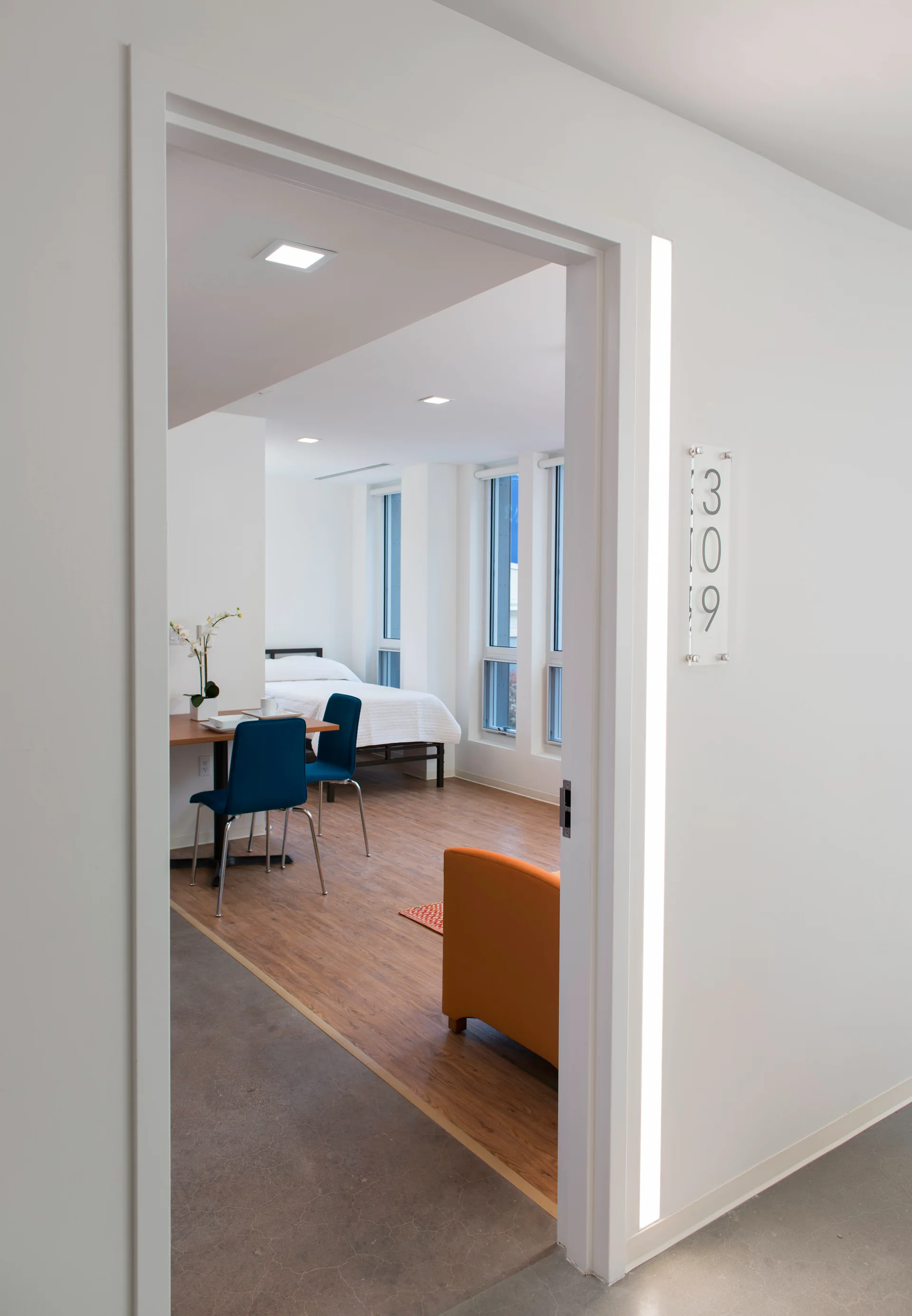 A view through a white door frame with a vertical light strip reveals a studio bedroom with a wood floor, bed, and dining table with blue chairs.