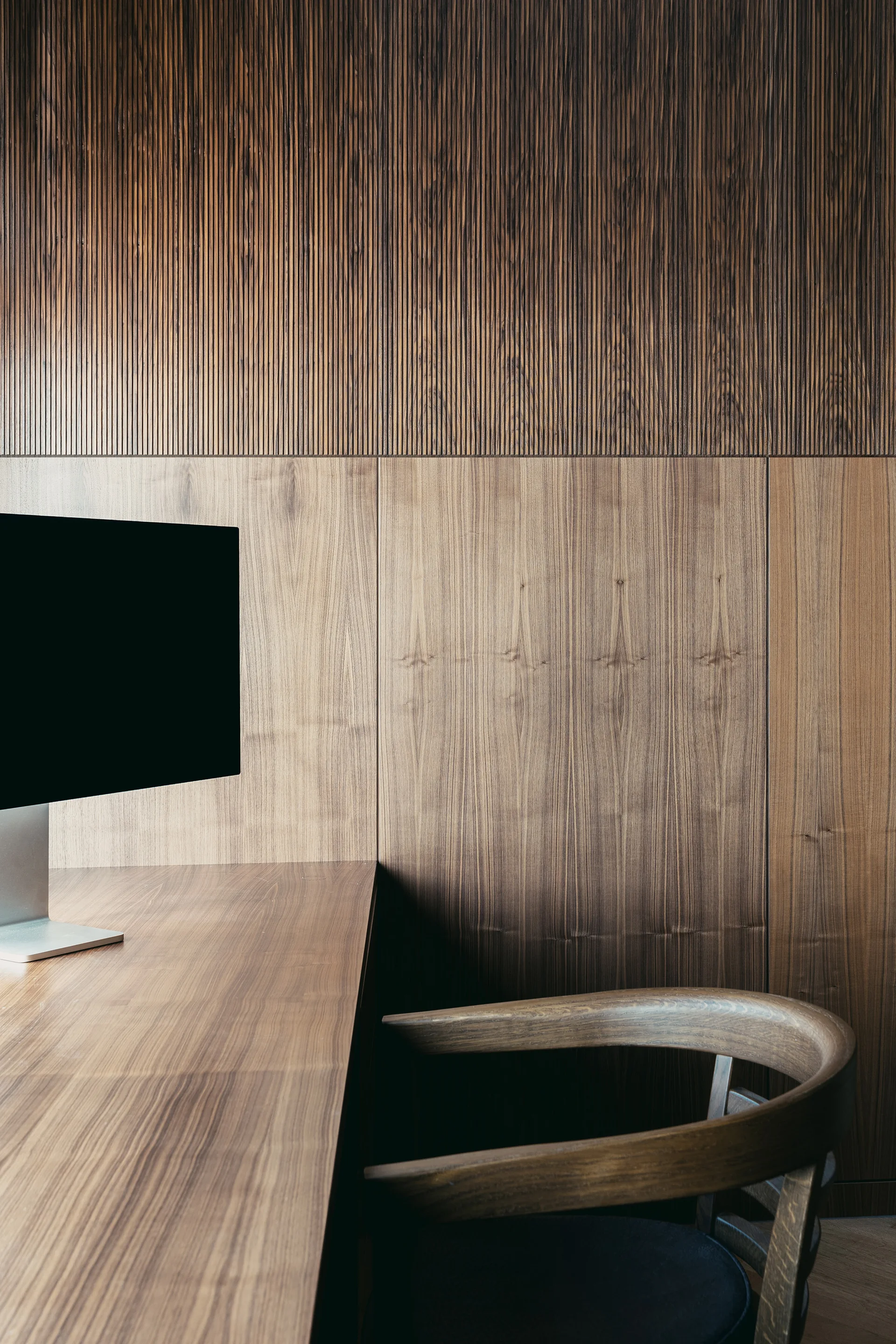 A minimalist home office features a curved wooden chair, a built-in wood desk with a monitor, and vertical wood-paneled walls.