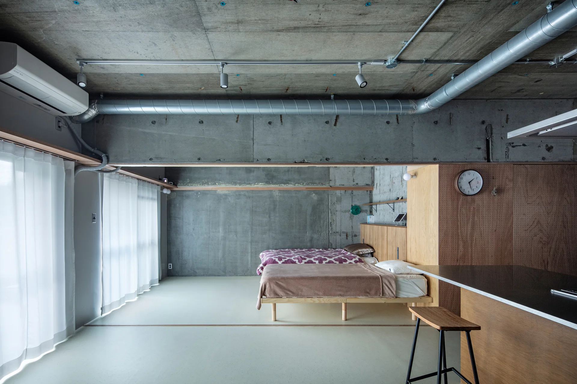 A minimalist bedroom features two twin beds set against a light wood paneled divider under a raw exposed concrete ceiling with silver ductwork.