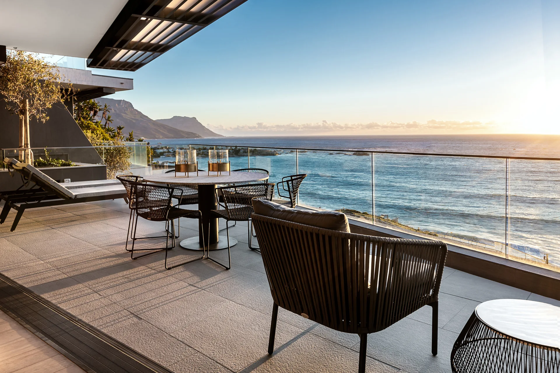 A modern outdoor terrace overlooking the ocean features a round stone dining table, woven black chairs, and a glass railing under a sunset sky.