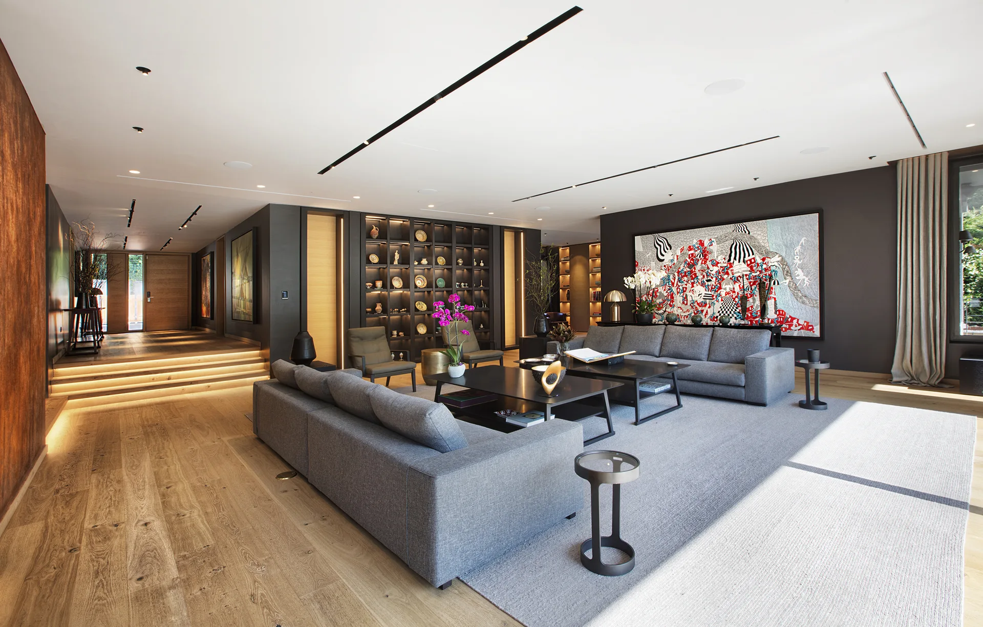A contemporary living room with grey sofas, black coffee tables, a large painting on a charcoal wall, and illuminated built-in shelving.