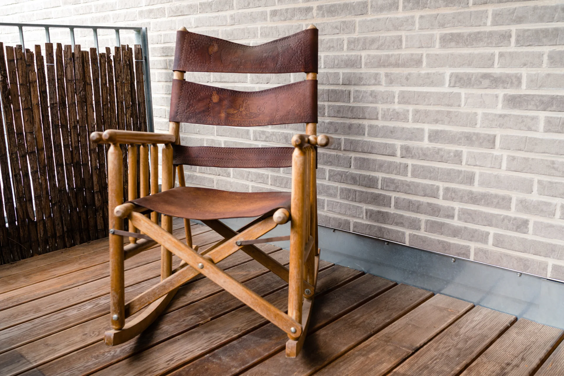 A rustic wooden rocking chair with etched brown leather straps sits on a wooden balcony deck against a grey brick wall.