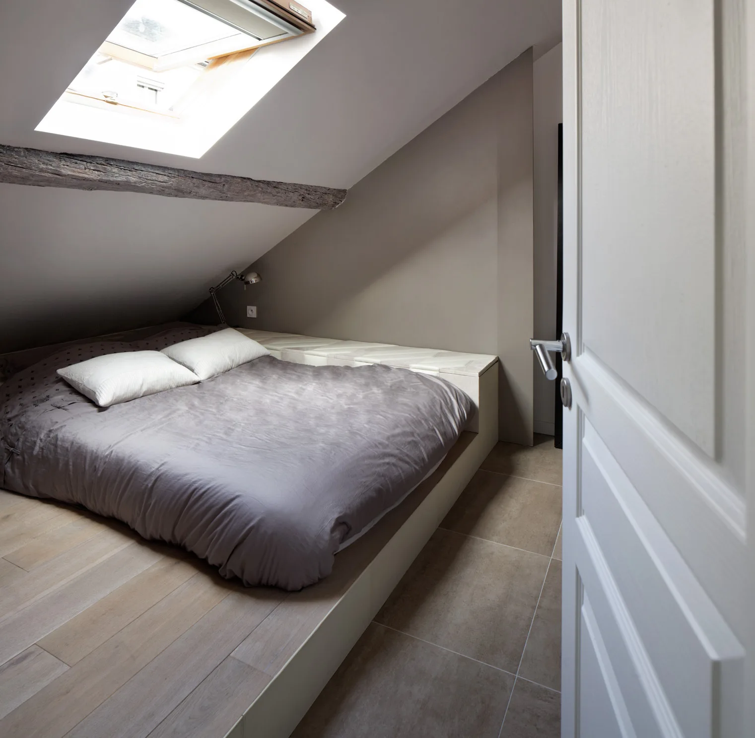 A low platform bed with grey bedding sits under a sloped attic ceiling featuring an exposed wood beam and a bright skylight.