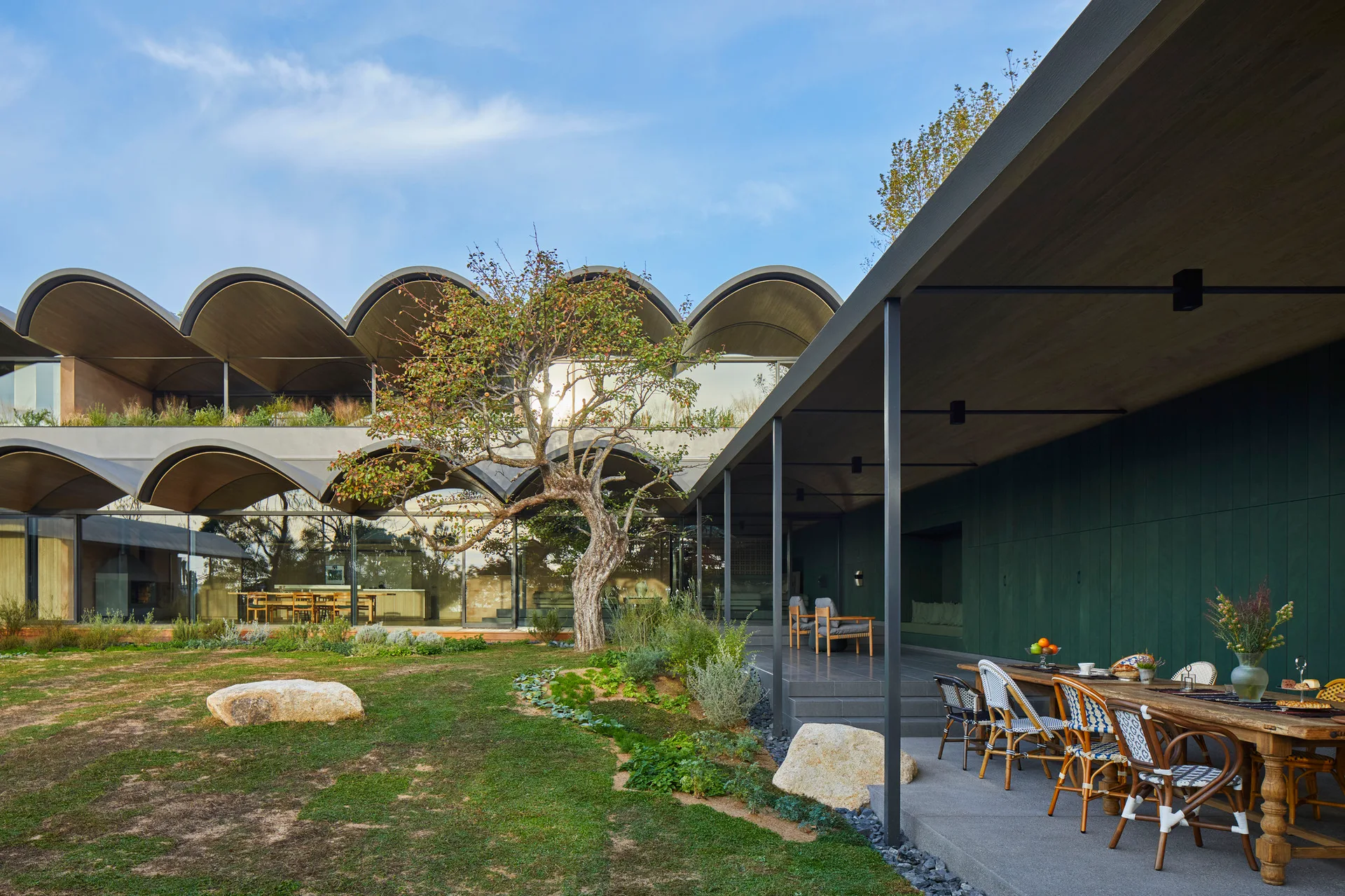 A covered stone patio features a rustic wood dining table with woven chairs next to a modern home with a repeating barrel vaulted roof and green walls.