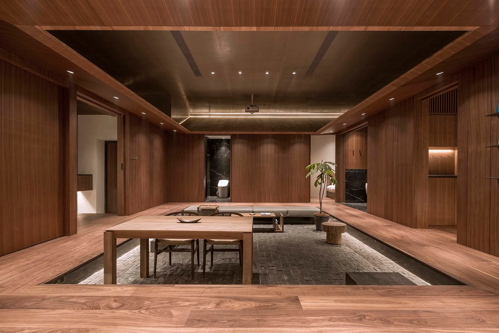 A minimalist room features warm wood paneled walls, a raised timber platform, and a sunken central seating area with grey furniture.