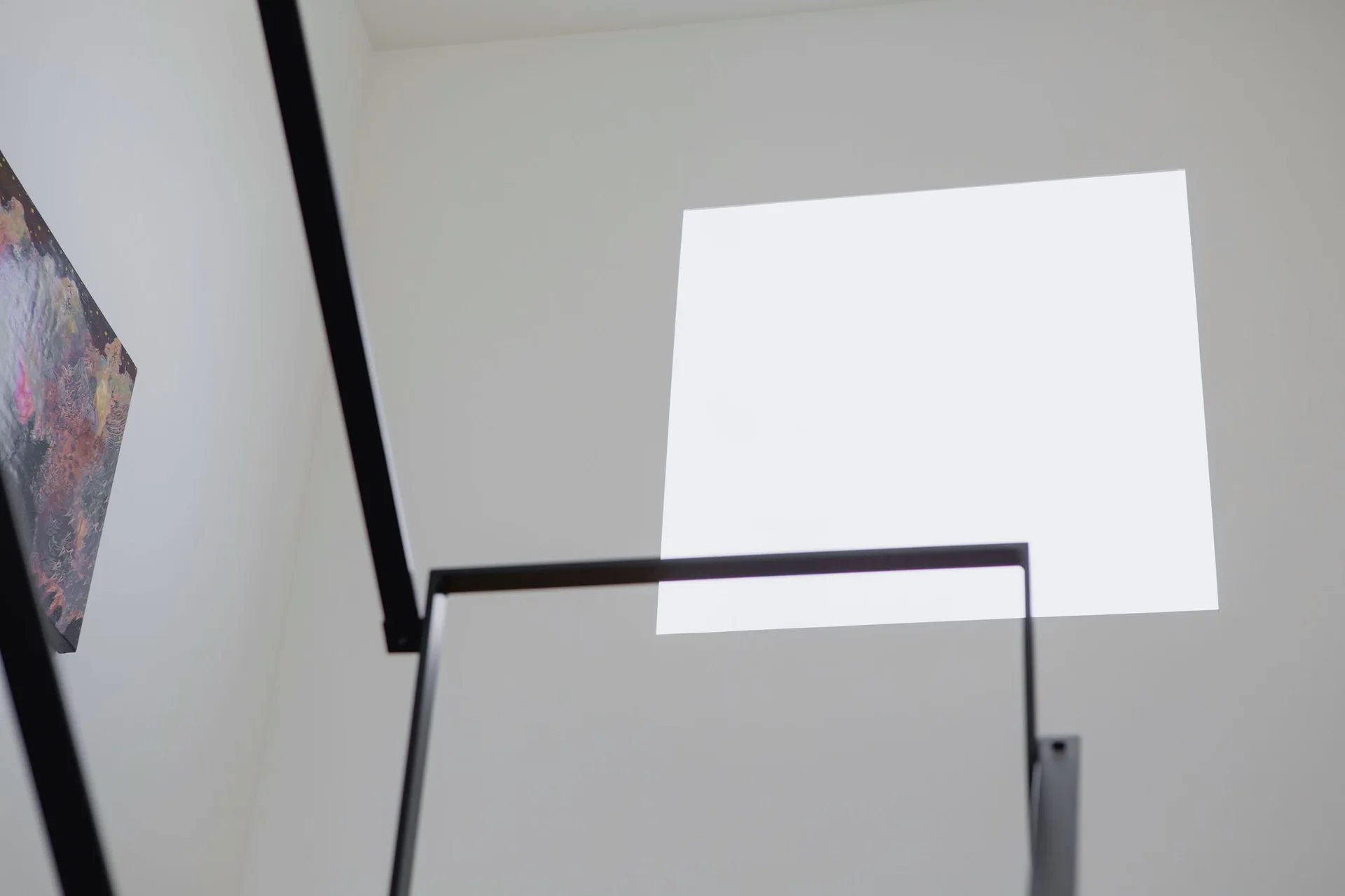 A thin matte black metal stair railing stands against clean white walls with a bright square window letting in daylight.