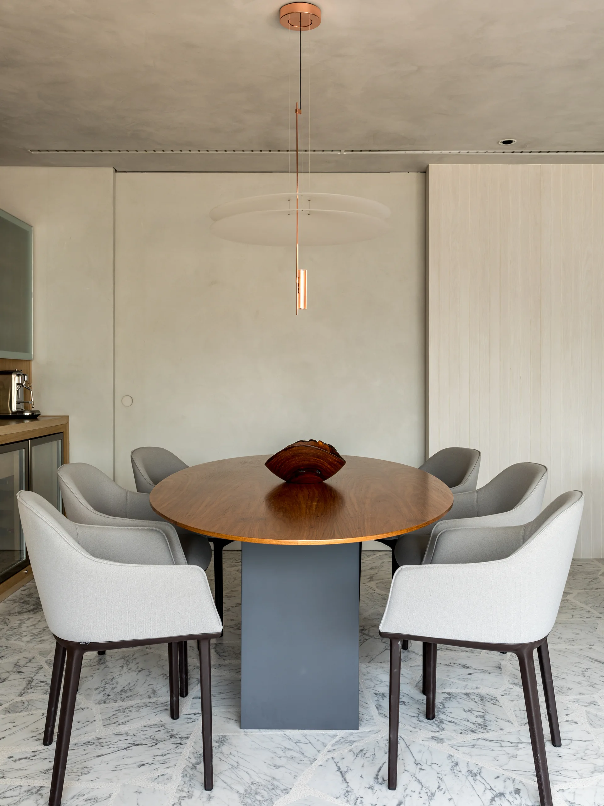 An oval walnut dining table surrounded by grey upholstered chairs sits on a veined marble floor beneath a minimalist copper pendant light.
