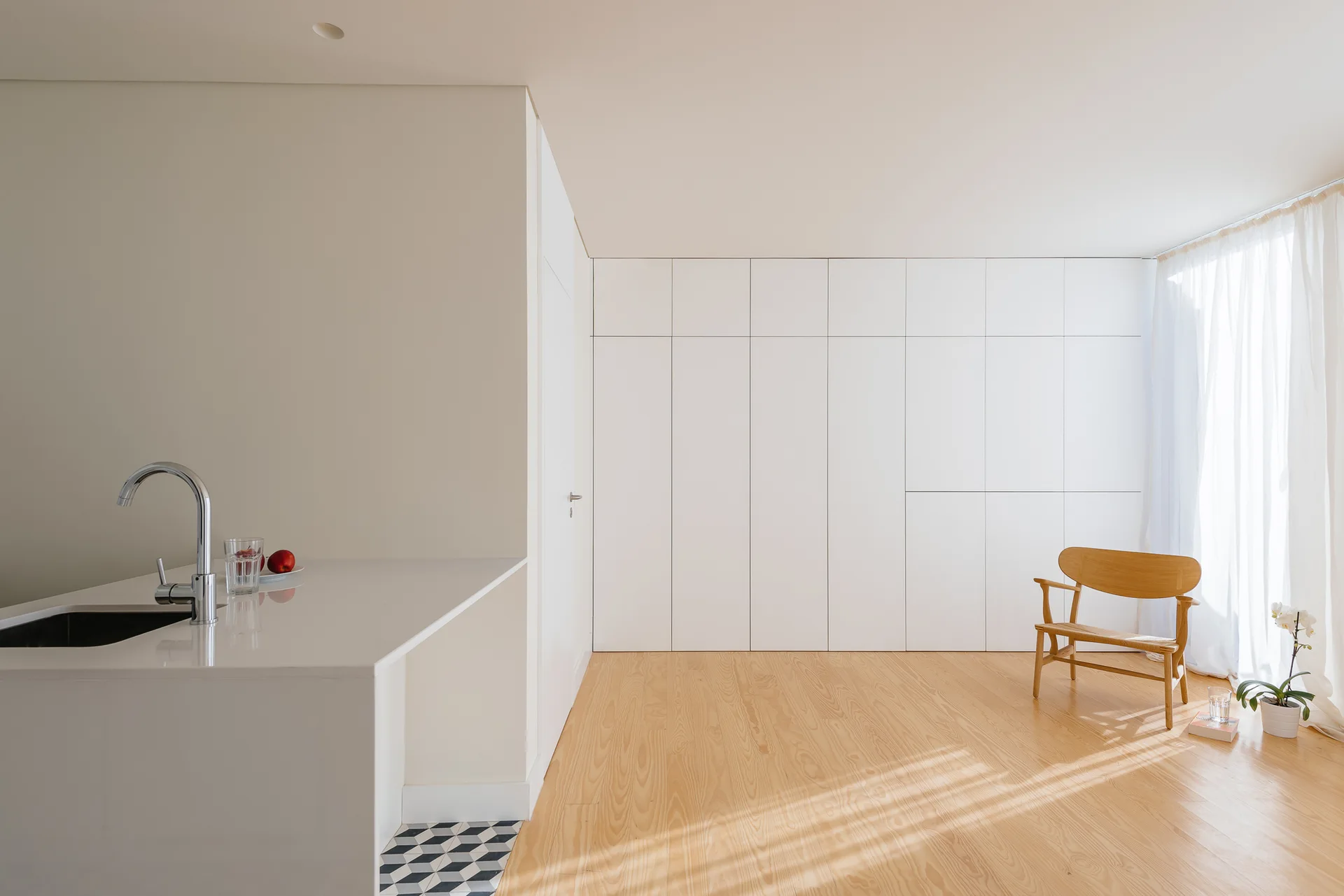 A minimalist kitchen space features a white counter with a chrome faucet, light wood flooring, and a large wall of handleless white cabinets.