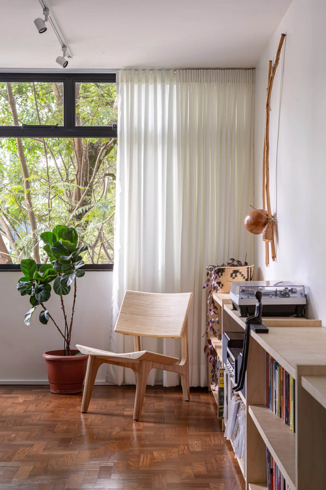 A molded plywood lounge chair sits on a parquet wood floor next to a low wooden bookshelf and a large window with white curtains.