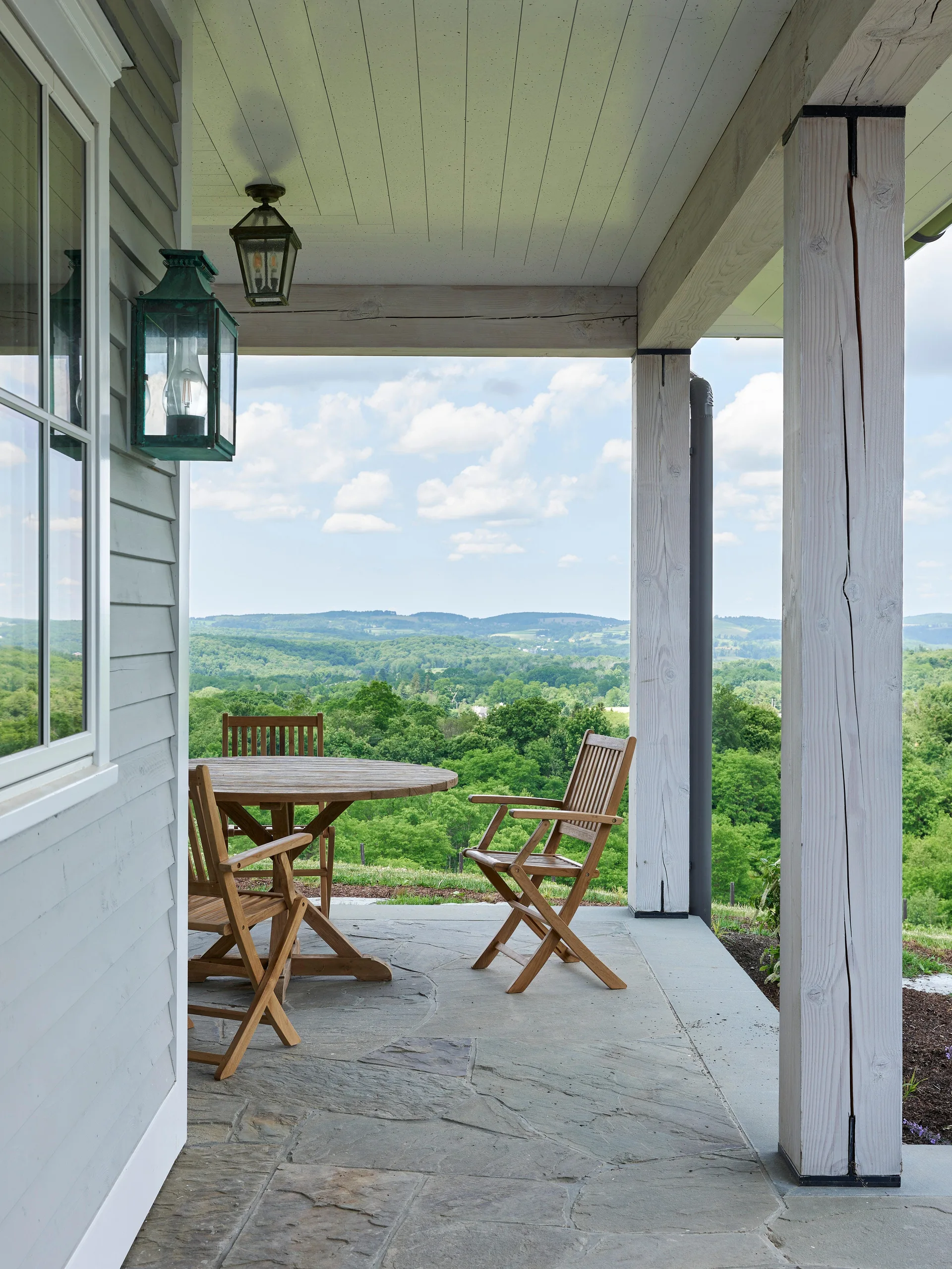 A covered farmhouse porch with wooden dining furniture, flagstone flooring, and thick columns looking out onto rolling green hills.
