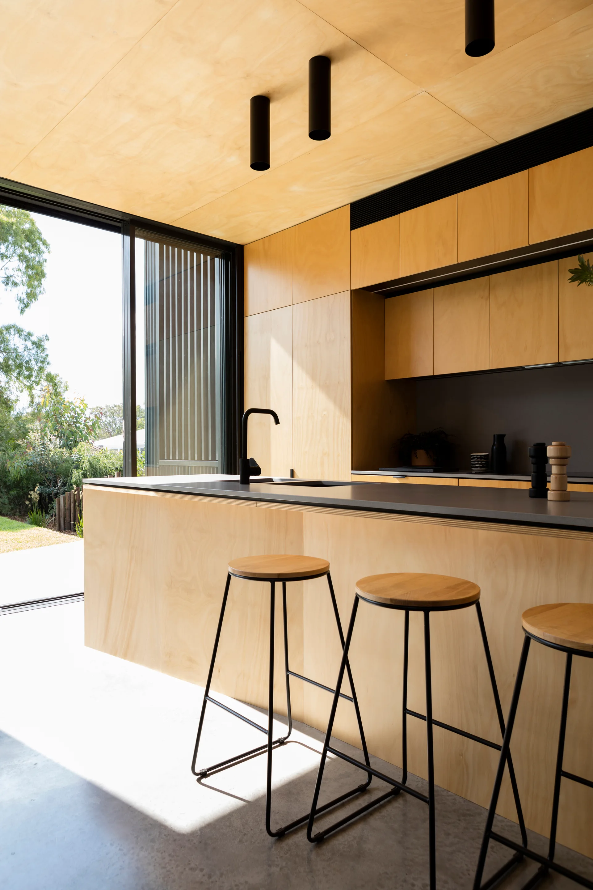 A modern kitchen featuring a plywood island and ceiling, concrete floors, three wooden bar stools, and a black faucet next to a sliding glass door.