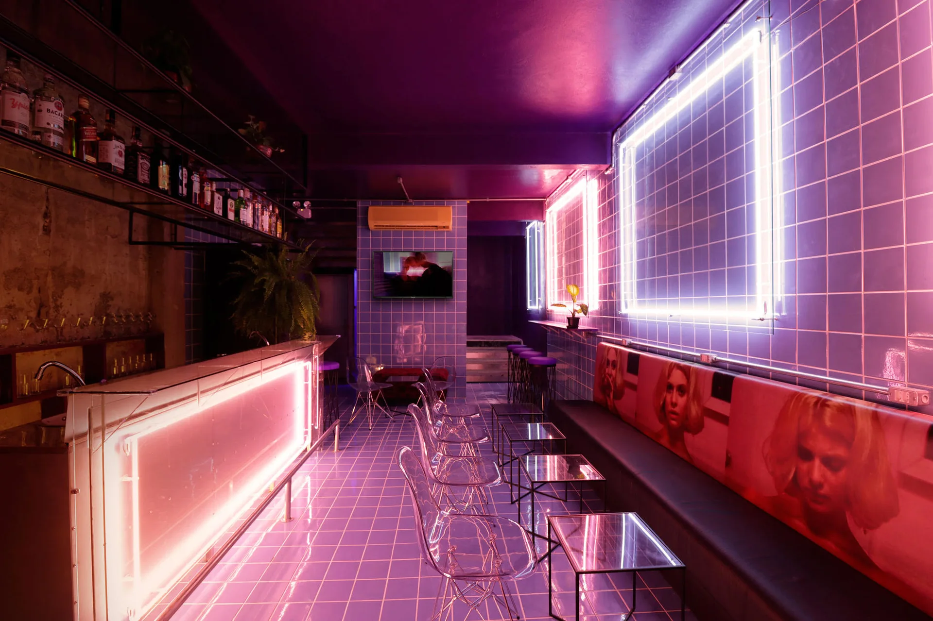 A commercial bar interior features saturated purple neon lighting, square grid tiles on the walls and floor, and transparent seating.