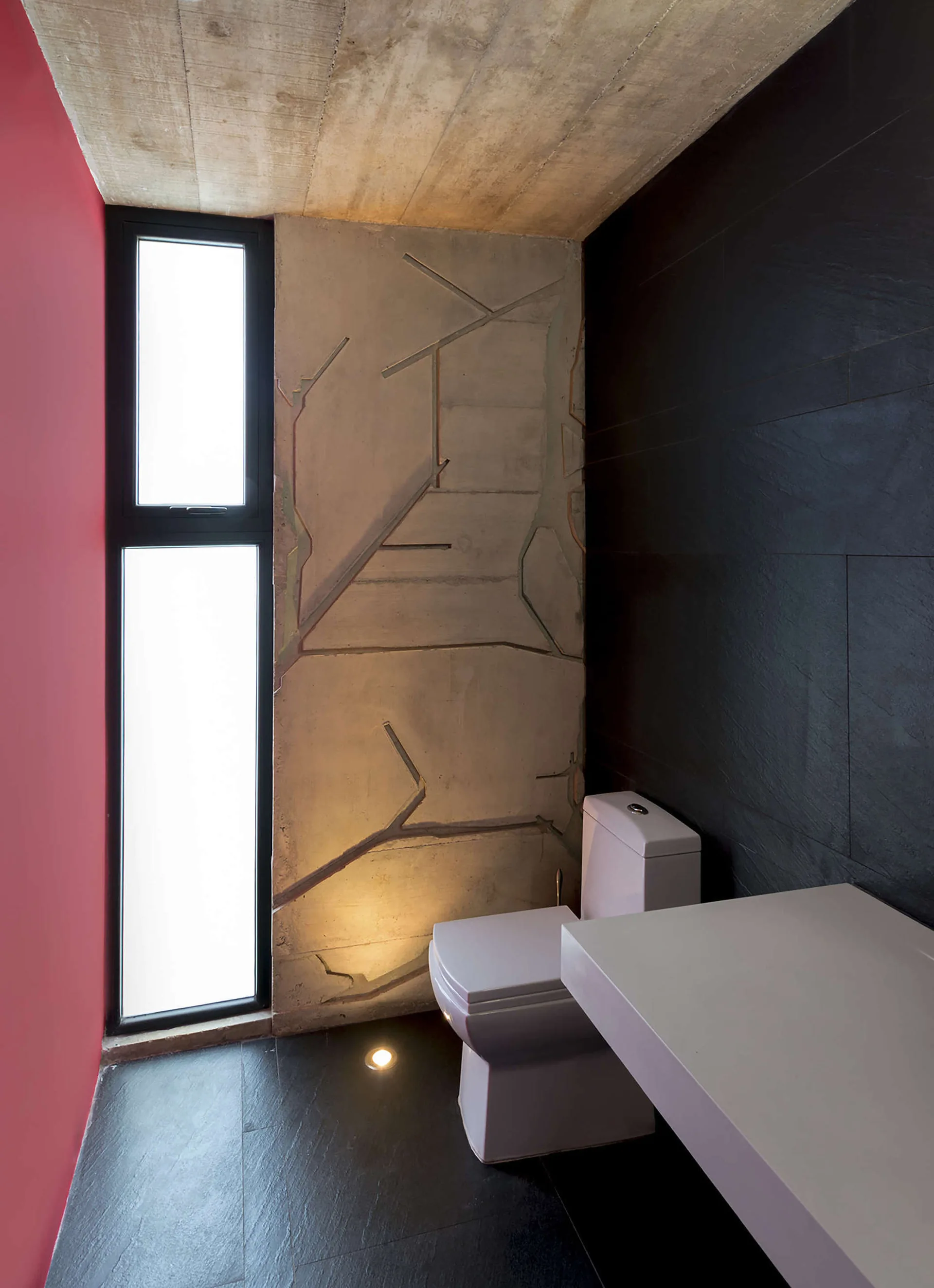 A minimalist powder room with a white toilet, a textured concrete accent wall, a tall narrow window, black floor tiles, and a red side wall.