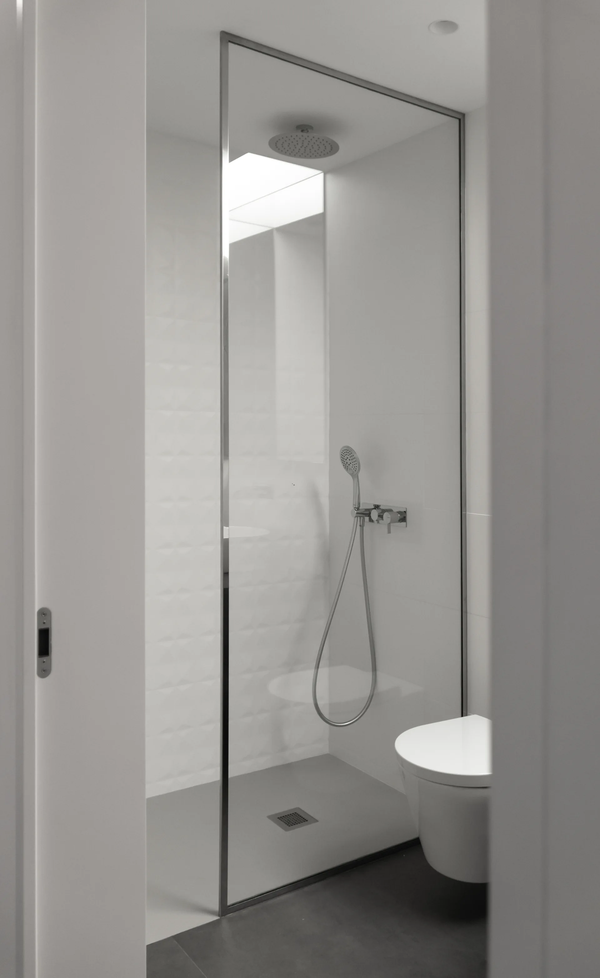 A modern bathroom viewed through a doorway featuring a frameless glass shower enclosure, a wall-mounted toilet, and white textured wall tiles.