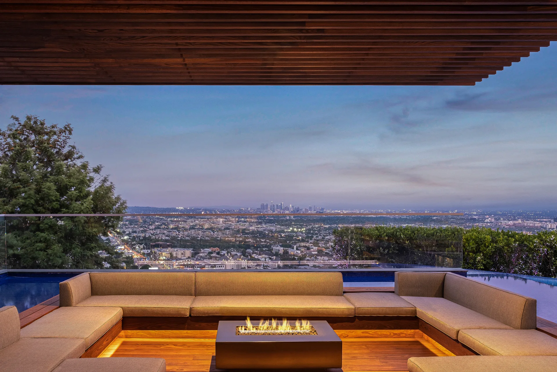 A U-shaped outdoor sectional sofa surrounds a lit square fire pit on a wood deck overlooking an infinity pool and city skyline at dusk.