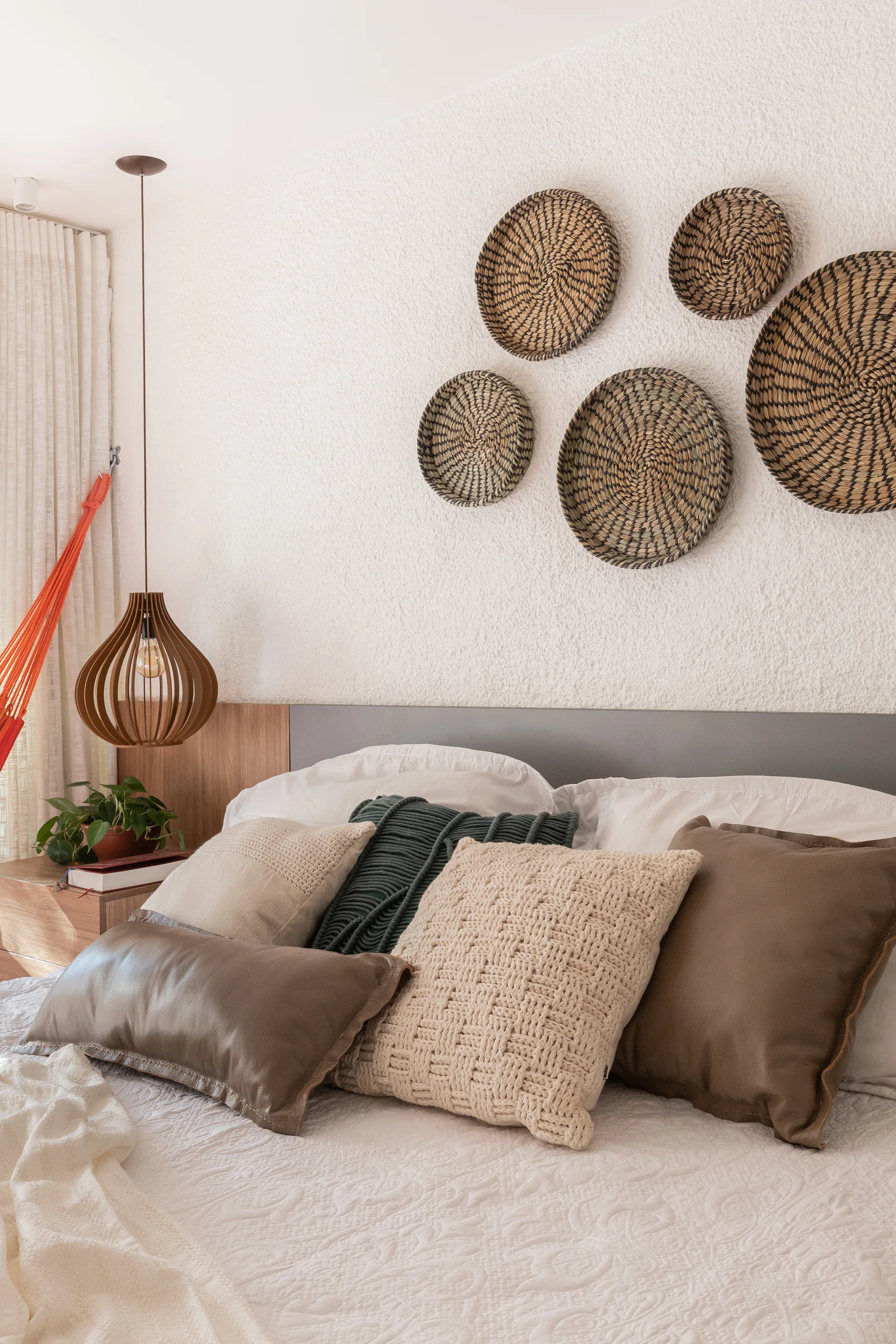 A bedroom featuring a collection of woven baskets displayed on a textured white wall above a bed layered with decorative knitted and leather pillows.