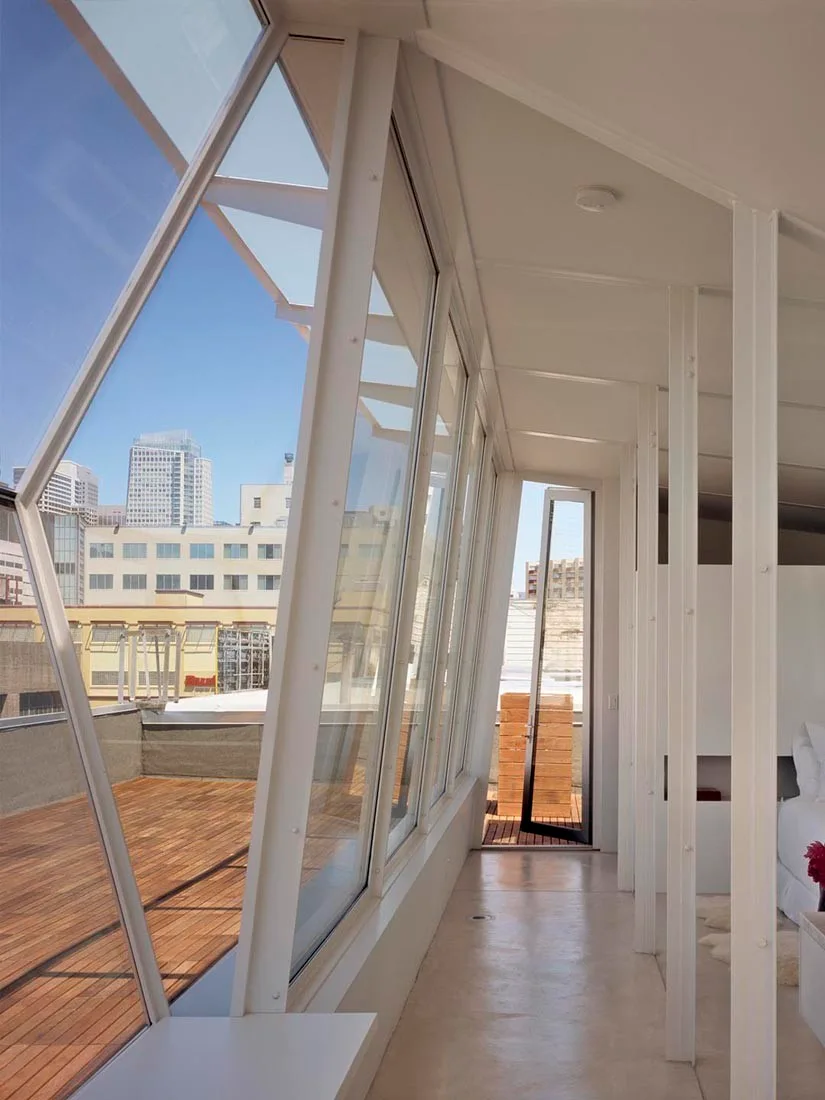 An interior hallway features a slanted glass window wall looking out onto a wooden deck terrace with city views.
