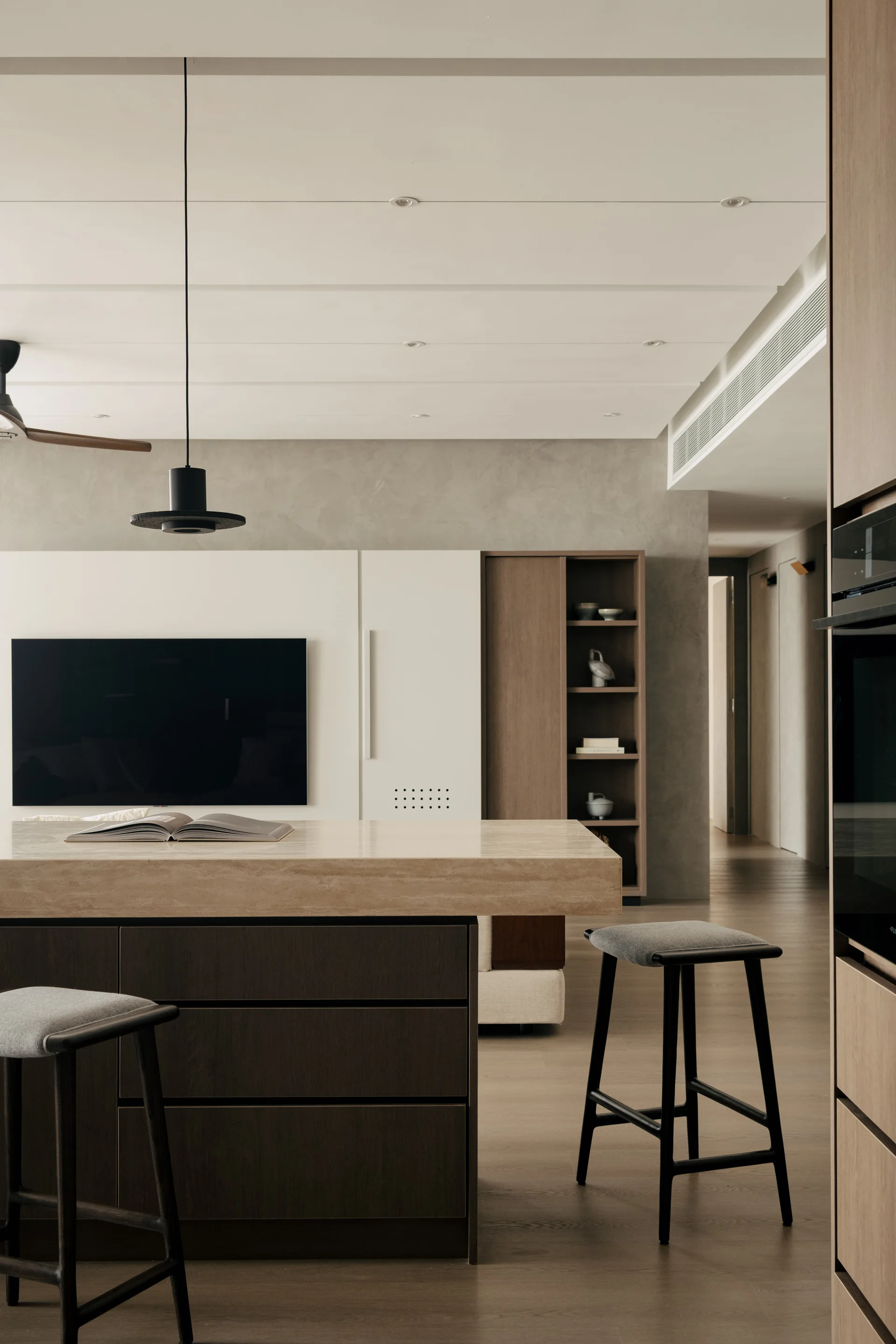 A minimalist kitchen features a stone-topped island with gray bar stools, dark wood cabinetry, and a flat-screen television on a textured plaster wall.