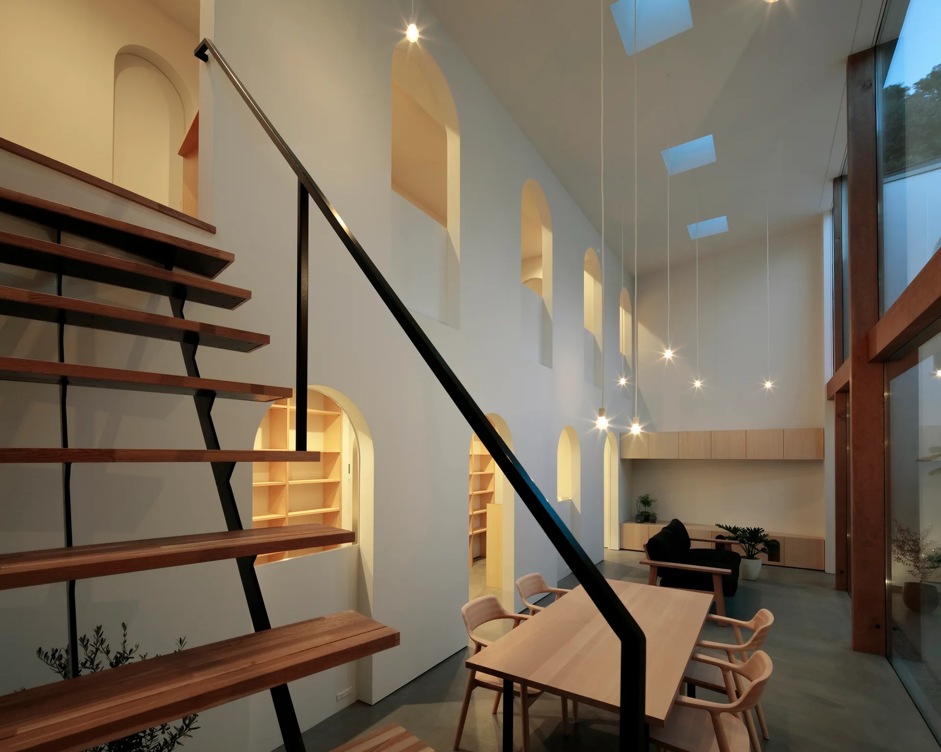 A minimalist dining space featuring a wood table, an open-riser staircase, and a double-height white wall punctuated by repeating arched openings.