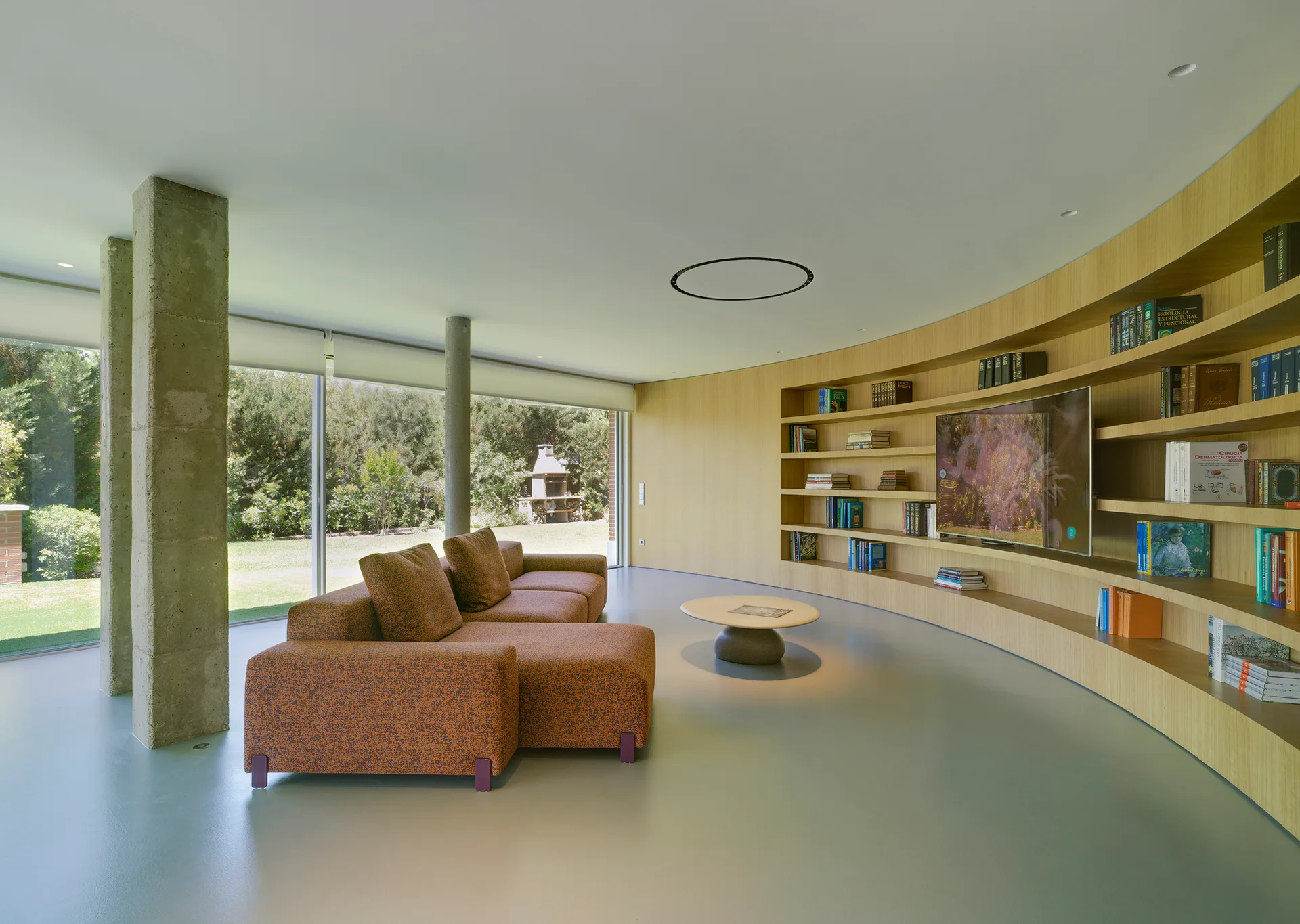 A contemporary living room features a curved wood bookcase wall, an orange modular sofa, exposed concrete pillars, and floor-to-ceiling windows.