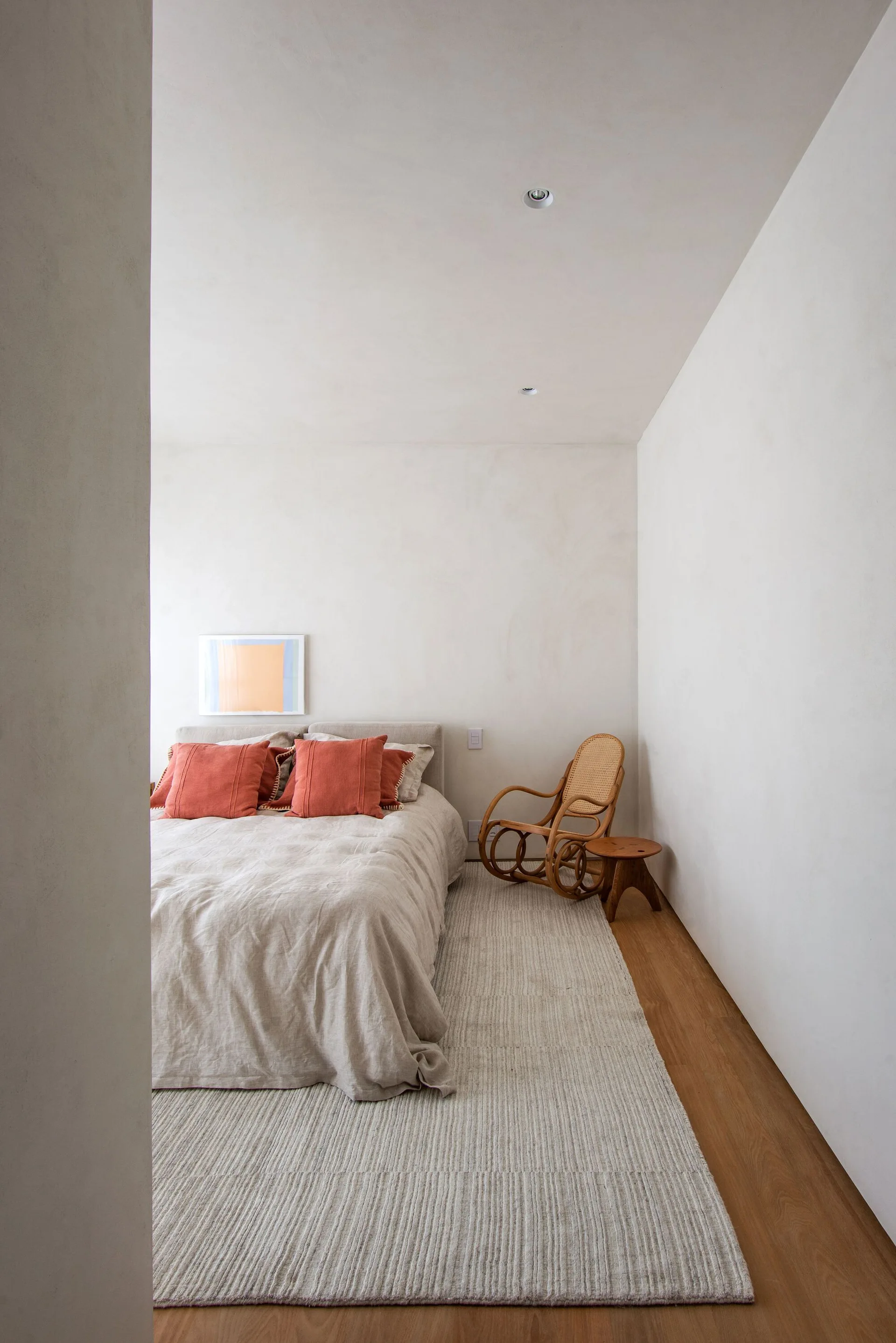 A minimalist bedroom featuring a bed with neutral linen sheets and terracotta pillows next to a classic wooden rocking chair on a textured rug.