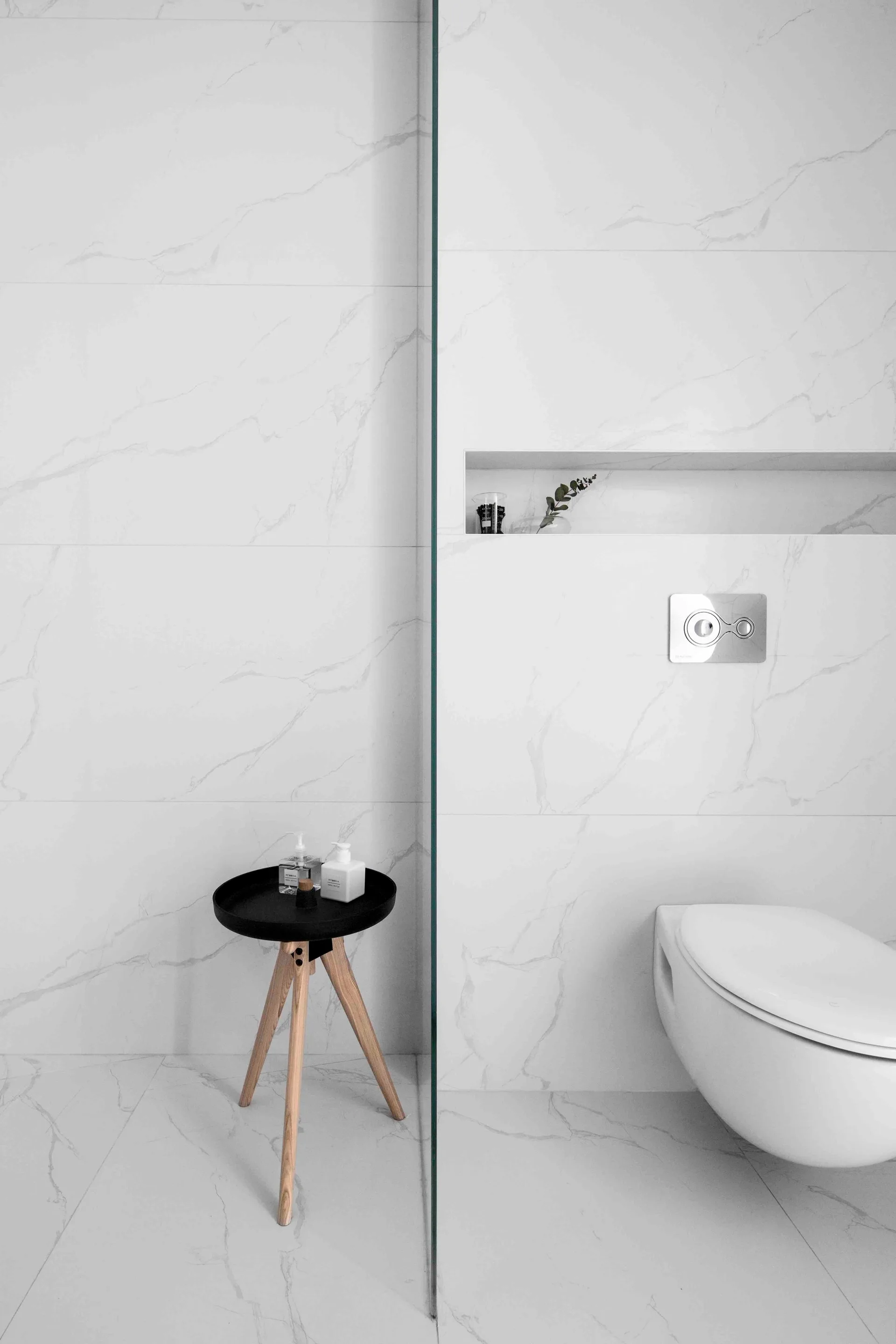 A minimalist bathroom features a wall-mounted toilet, a glass shower partition, marble-patterned tiles, and a small black tripod side table.