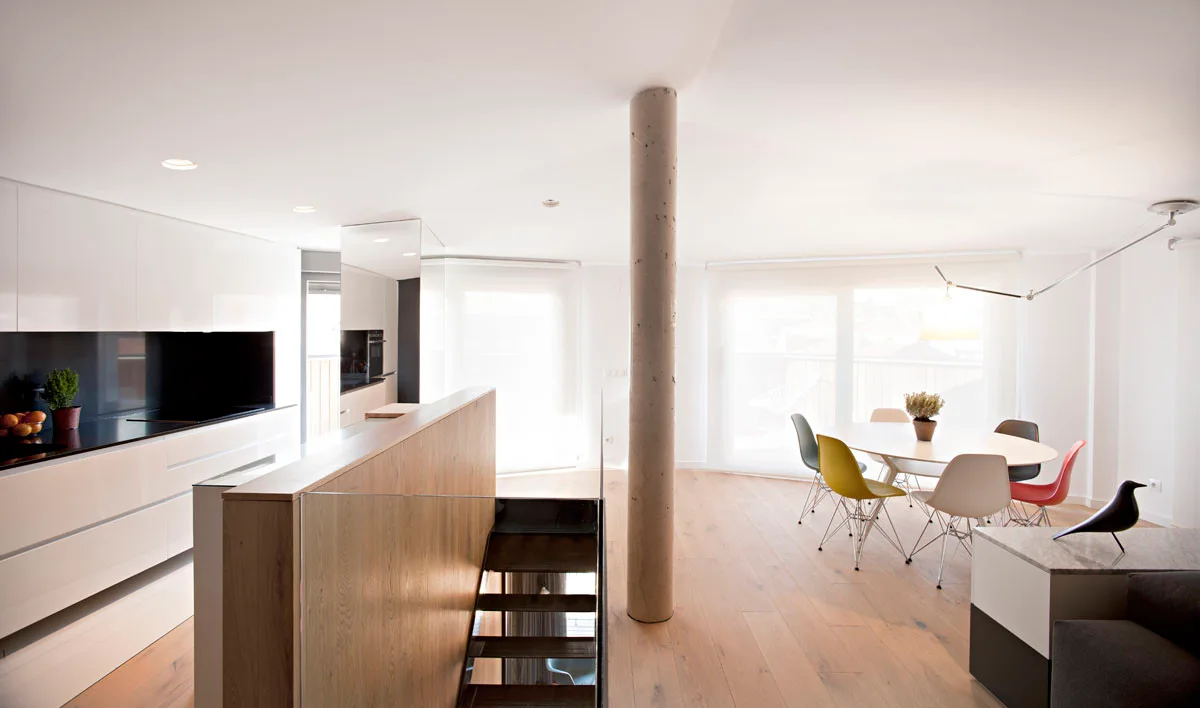 A modern open-plan kitchen and dining area with a white oval table, colorful chairs, a central concrete column, and a wooden staircase.