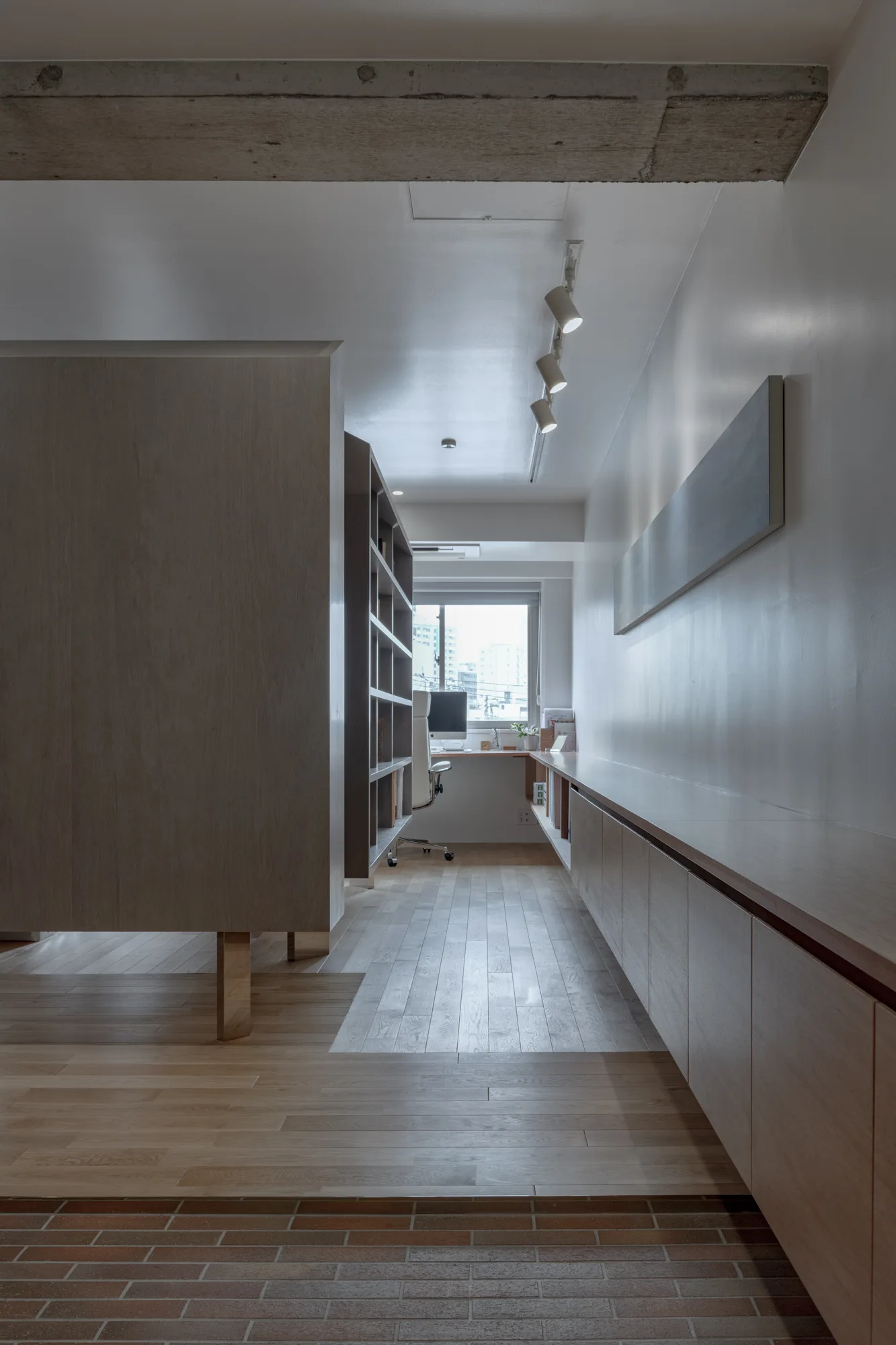 A narrow minimalist home office features a long low wooden cabinet, a wood room divider, track lighting, and a desk under a far window.