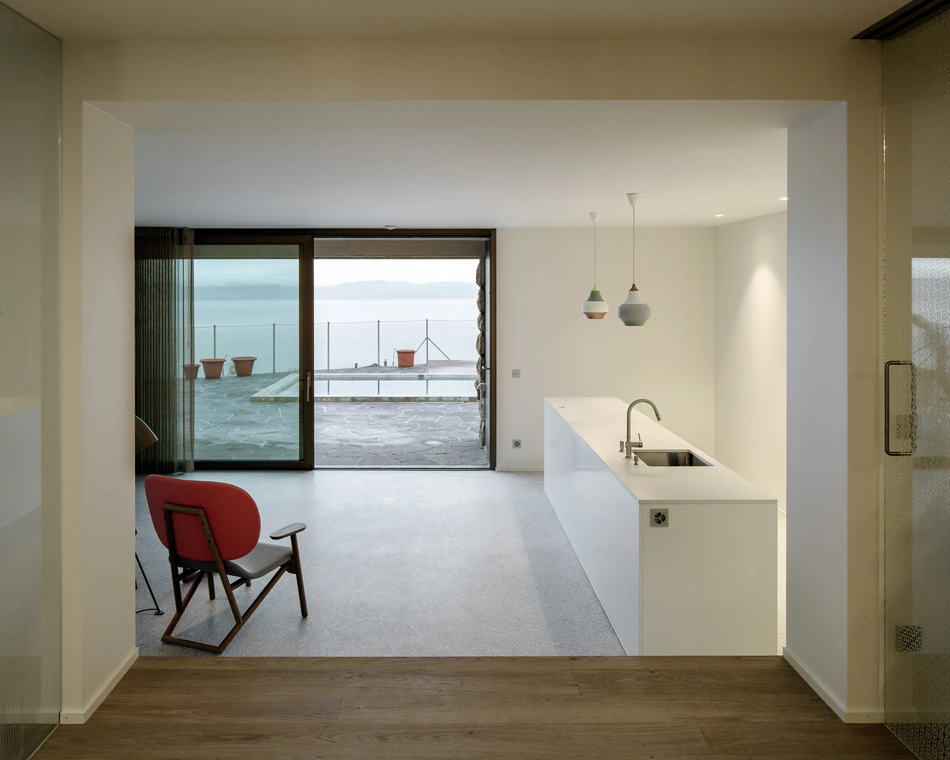 A minimalist open-plan kitchen with a white kitchen island and a red chair faces sliding glass doors that open to a pool terrace and coastal view.