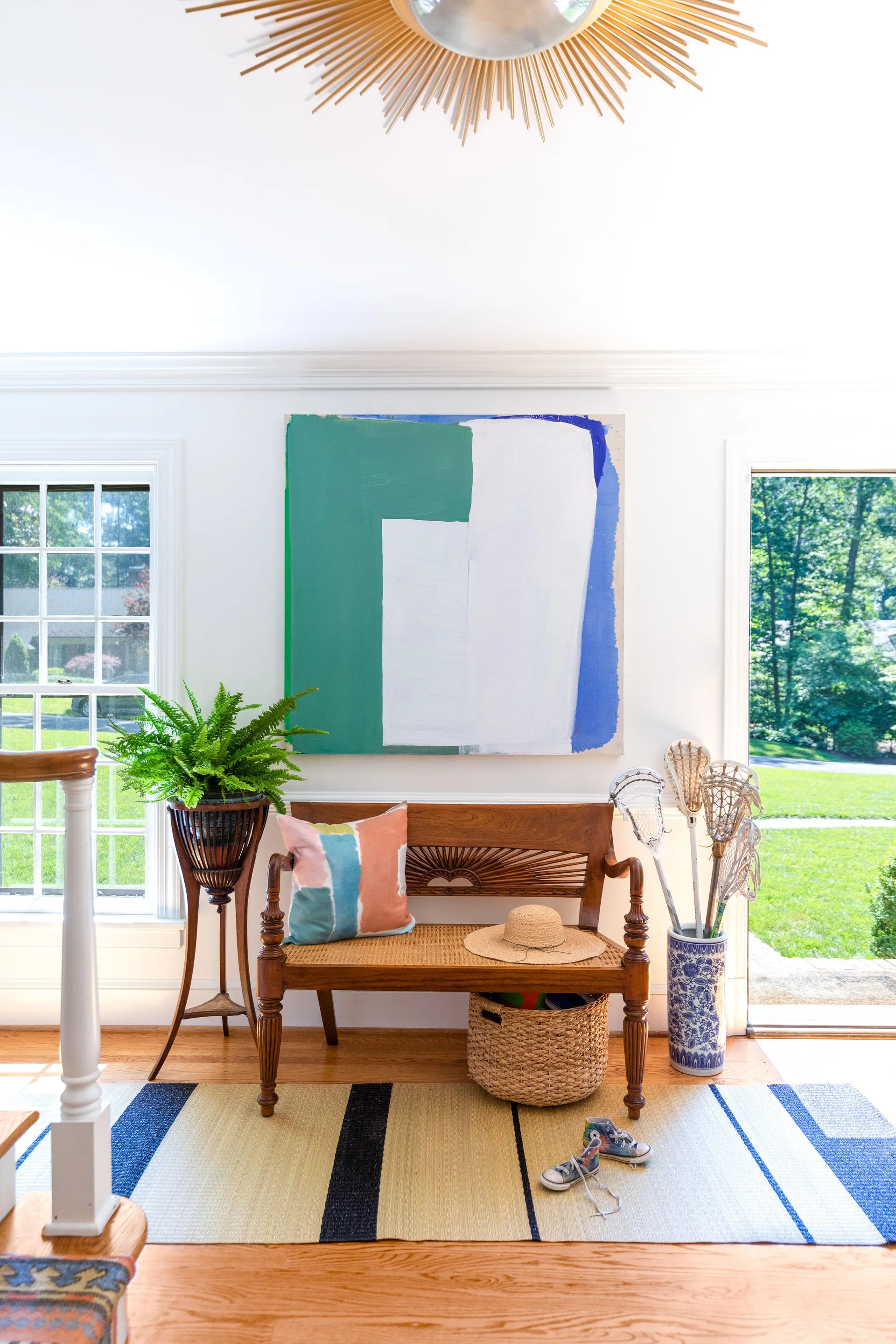 An entryway features a wooden bench with a cane seat beneath a green and blue abstract painting, flanked by a potted fern and sports equipment.
