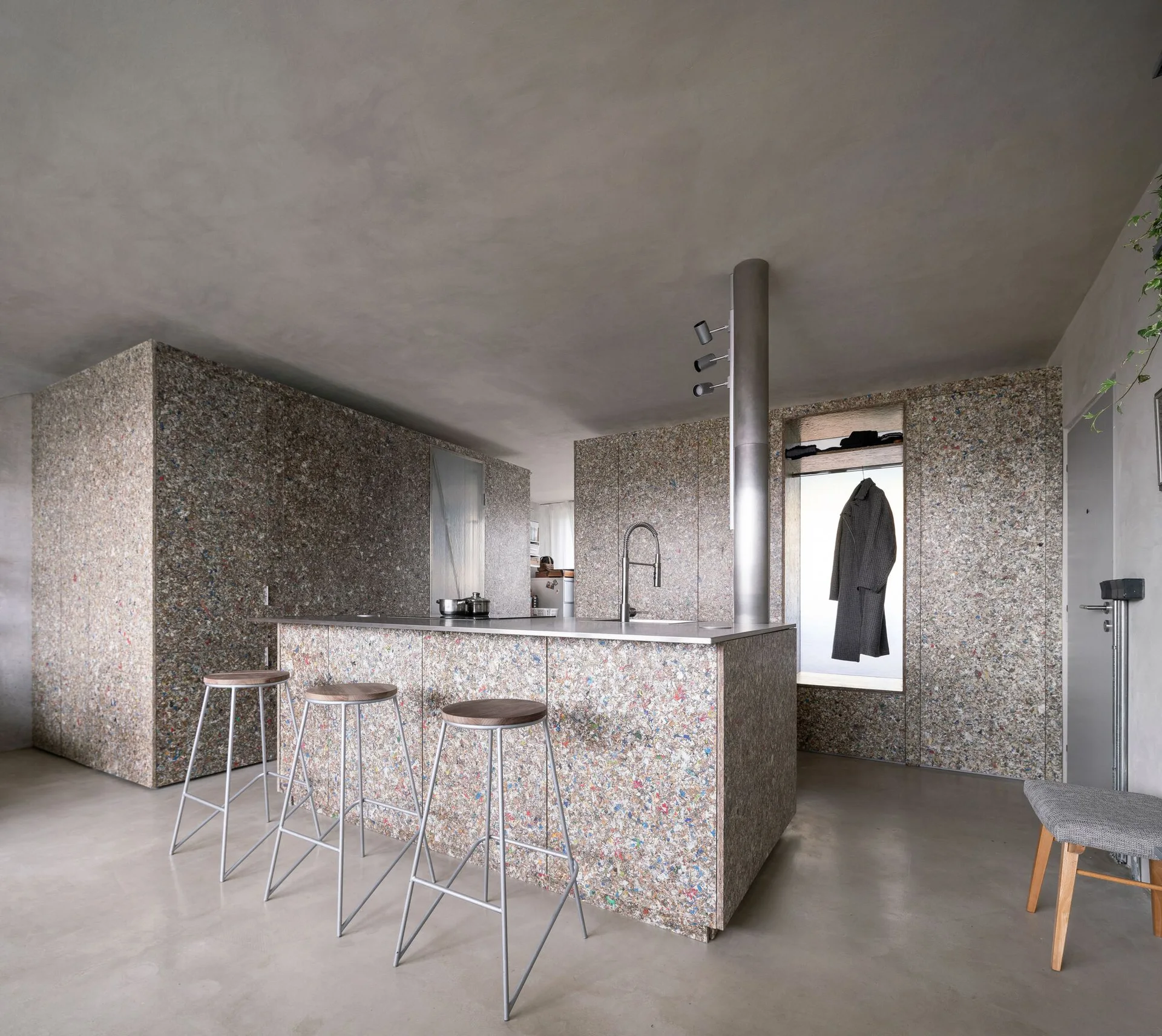 A minimalist kitchen features a central island and cabinetry clad in speckled multi-colored composite panels, paired with tall metal bar stools.