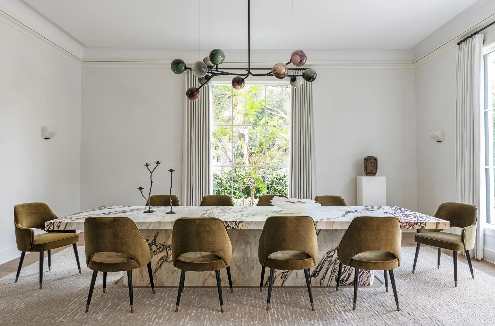 A large veined marble dining table is surrounded by olive green velvet chairs under a modern branching glass orb chandelier.