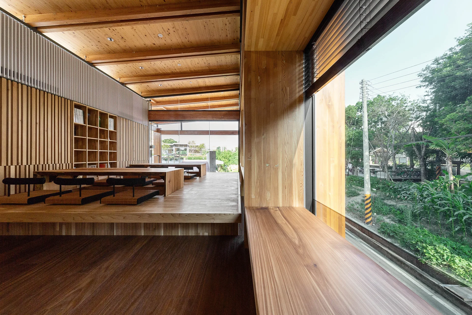 A minimalist wooden interior features a raised seating platform with low tables, vertical wood slat walls, and a large window looking outside.