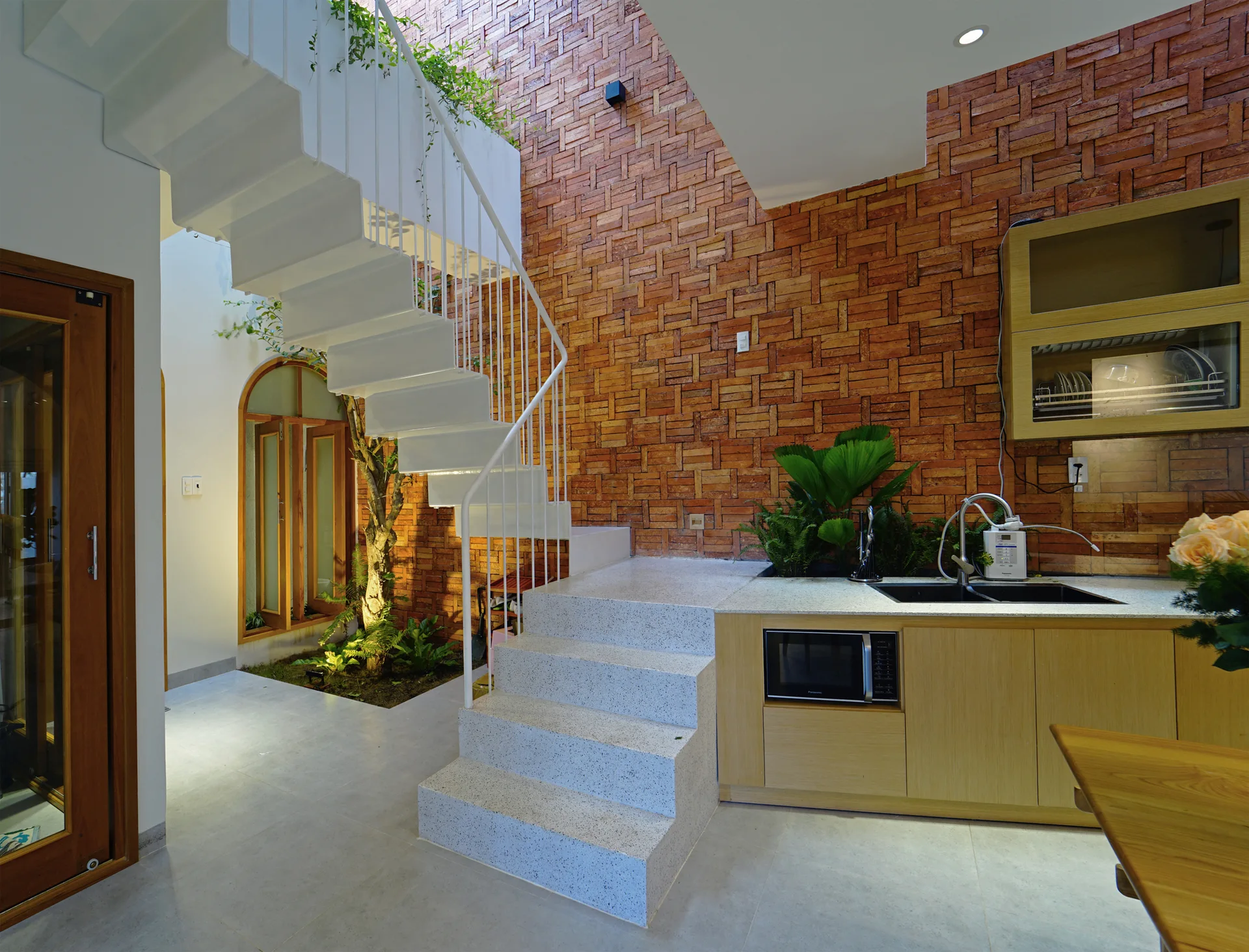 A modern kitchen features light wood cabinets, a black sink, a textured red brick wall, and a white metal staircase rising over a light stone landing.