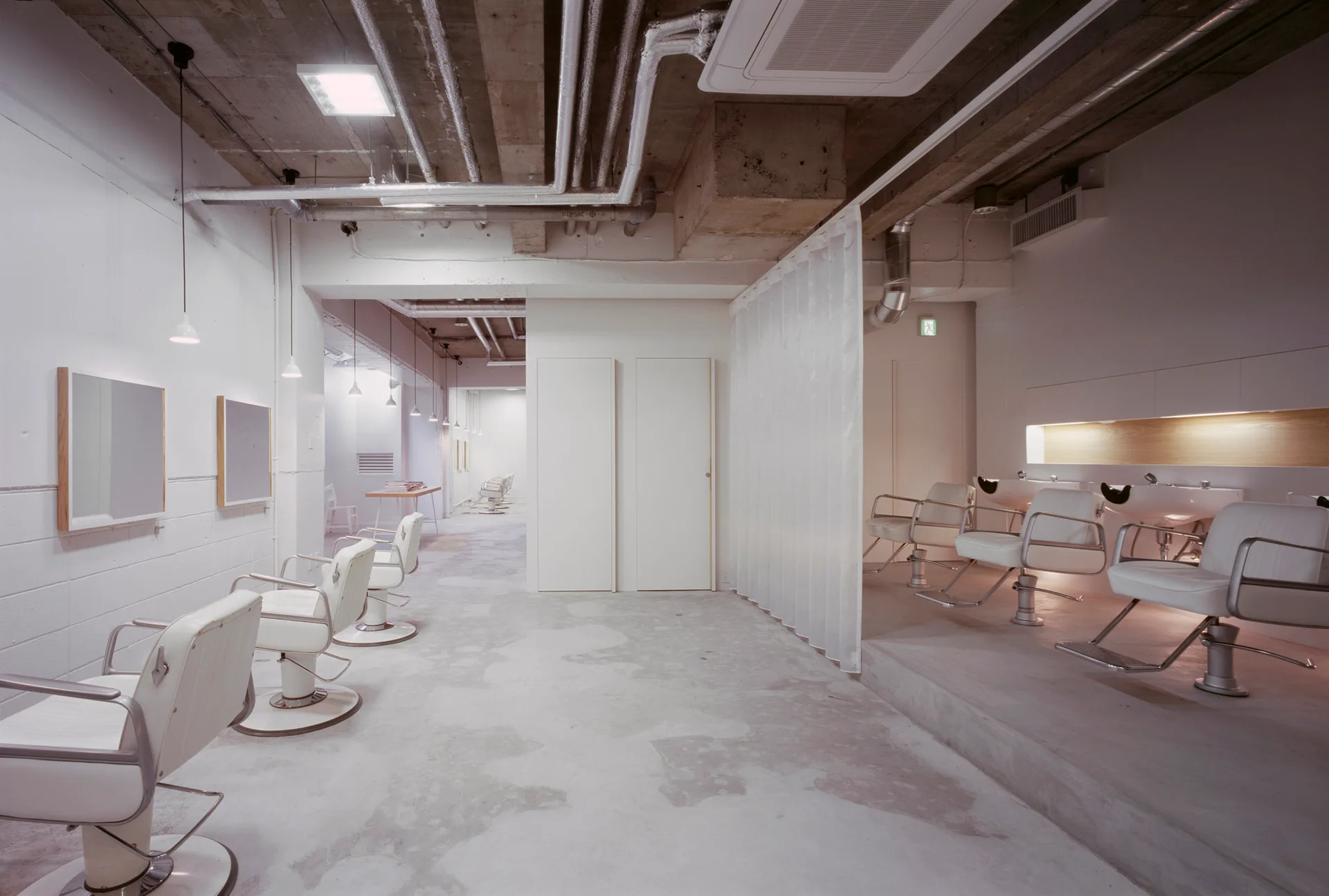 A minimalist white salon features rows of styling chairs, shampoo sinks on a raised platform, and an industrial exposed concrete ceiling.