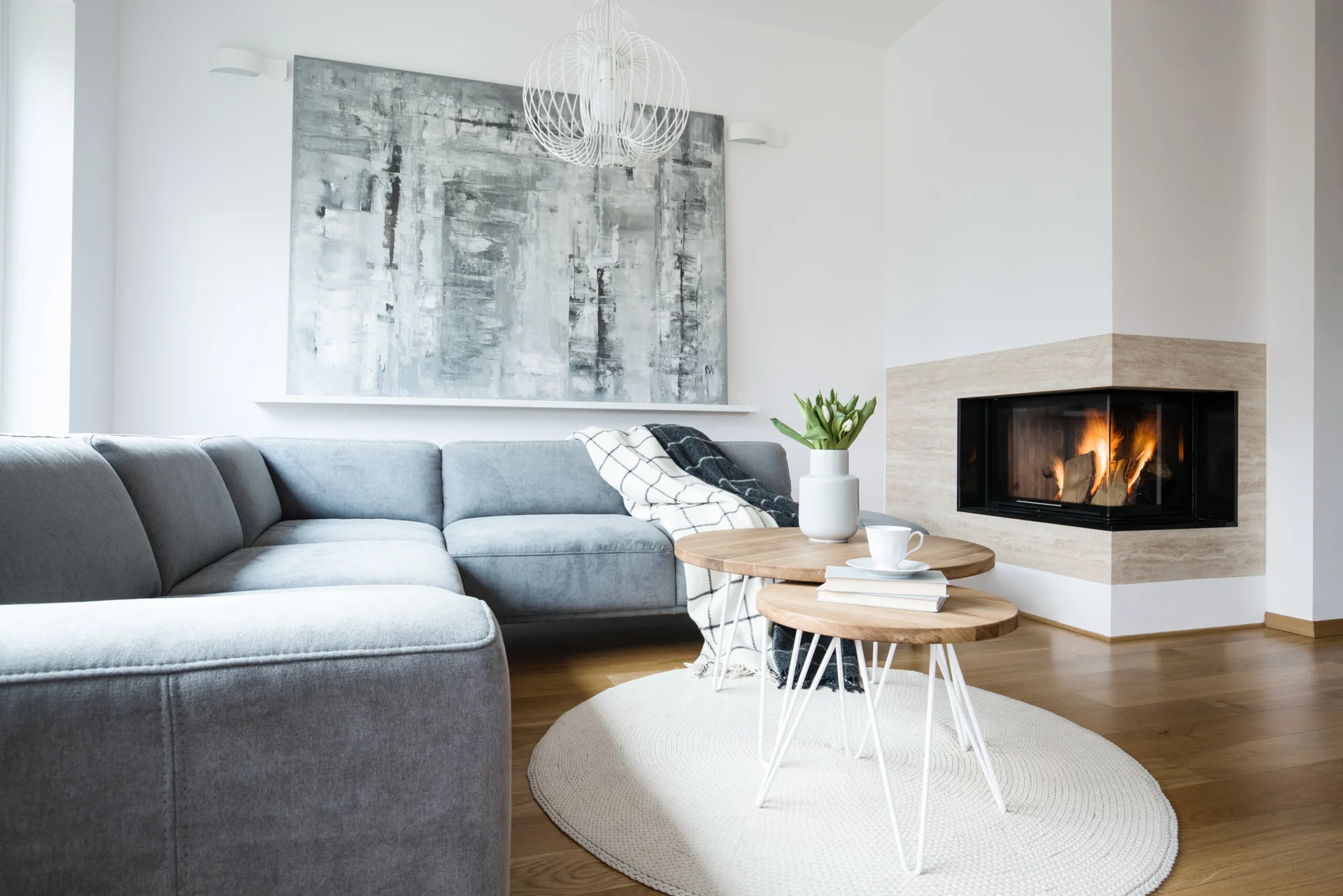 A modern living room features a grey sectional sofa, nested wooden coffee tables on a round rug, and a burning corner fireplace.