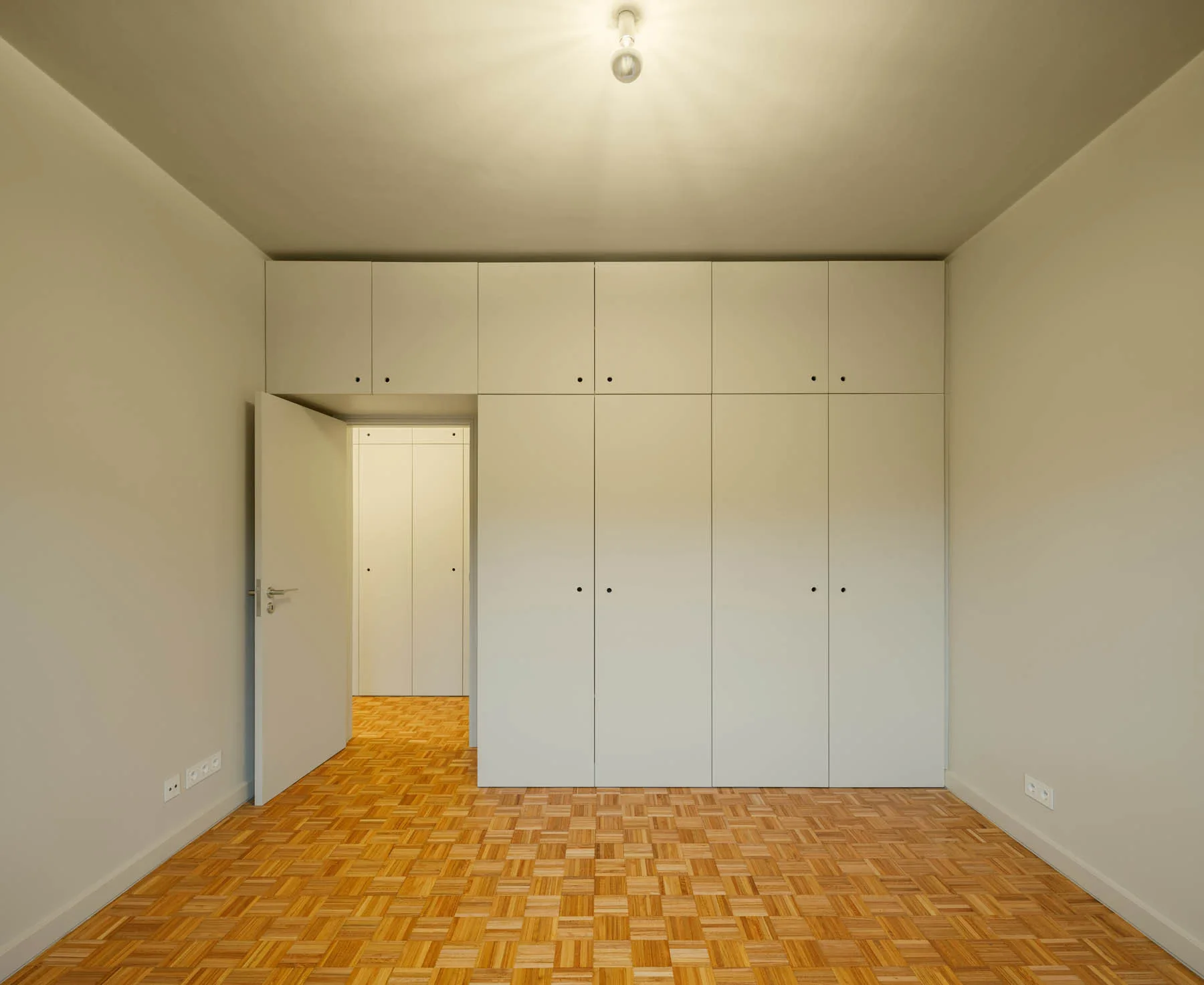 A minimalist room featuring white floor-to-ceiling built-in wardrobes and a patterned wooden parquet floor next to an open doorway.