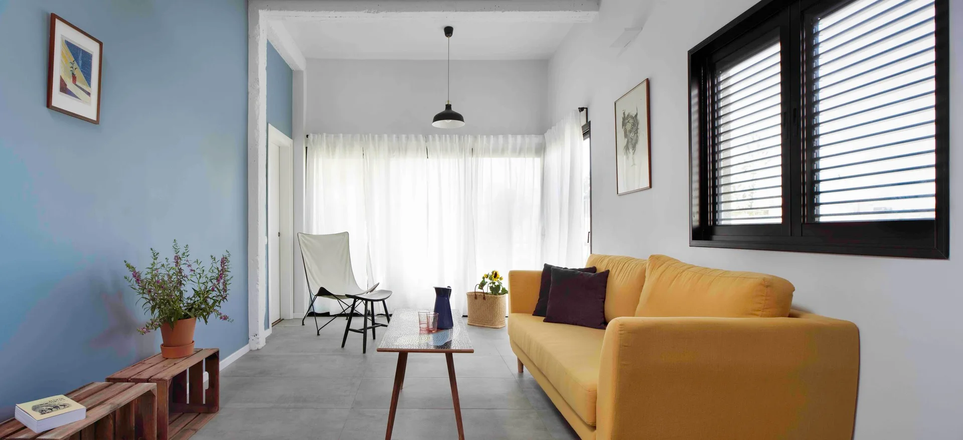 A modern living room featuring a bright yellow sofa, a slate blue accent wall, a white butterfly chair, and floor-to-ceiling sheer white curtains.