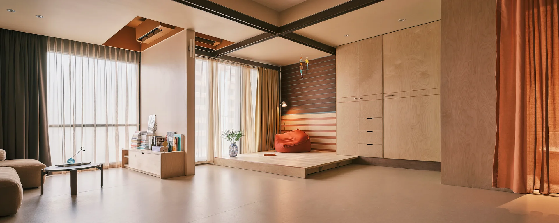 A modern living room features a raised wooden platform with an orange bean bag chair, built-in plywood cabinetry, and floor-to-ceiling sheer curtains.