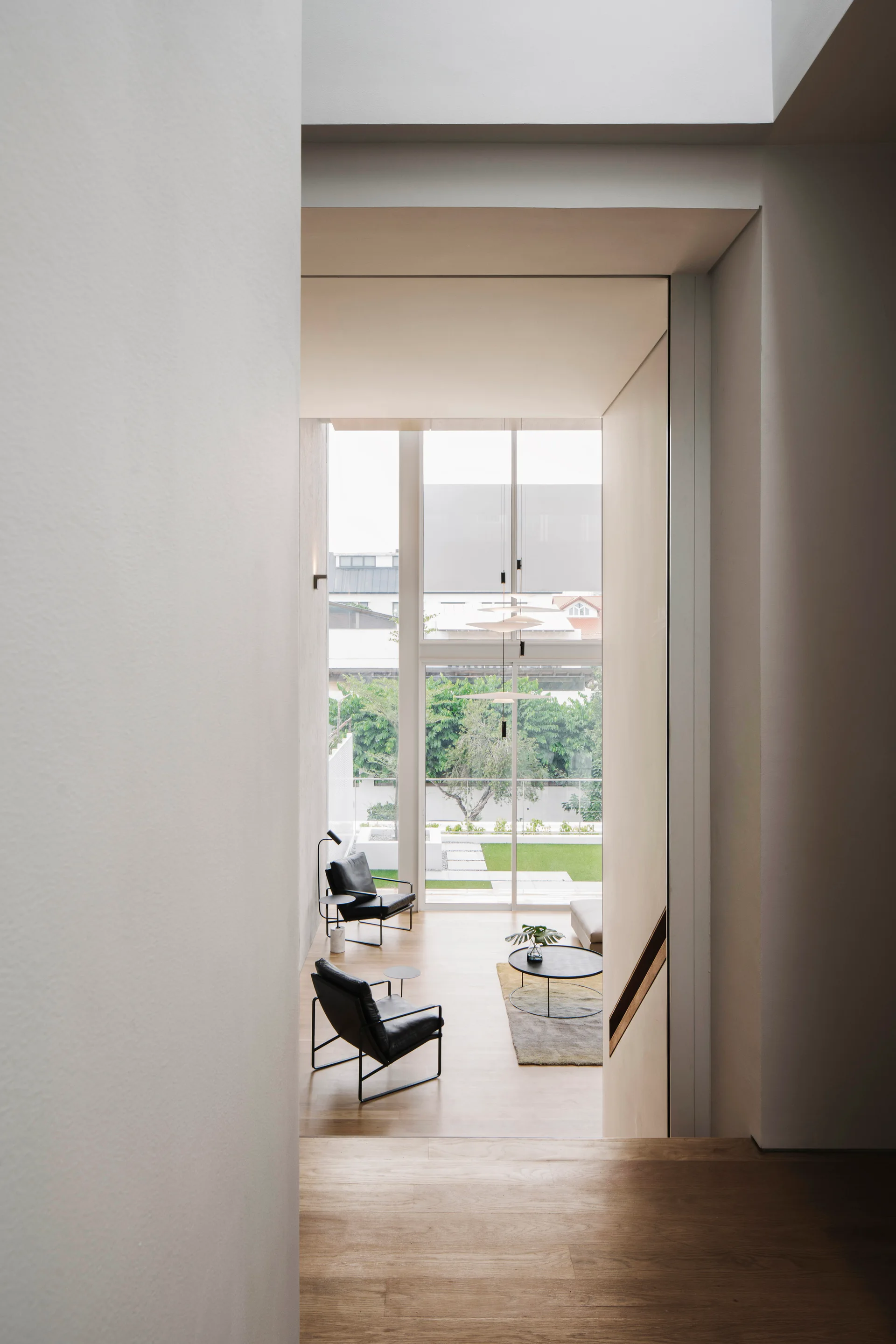 A high-angle view looking down wooden stairs into a minimalist living room with black leather armchairs, a round coffee table, and large glass windows.