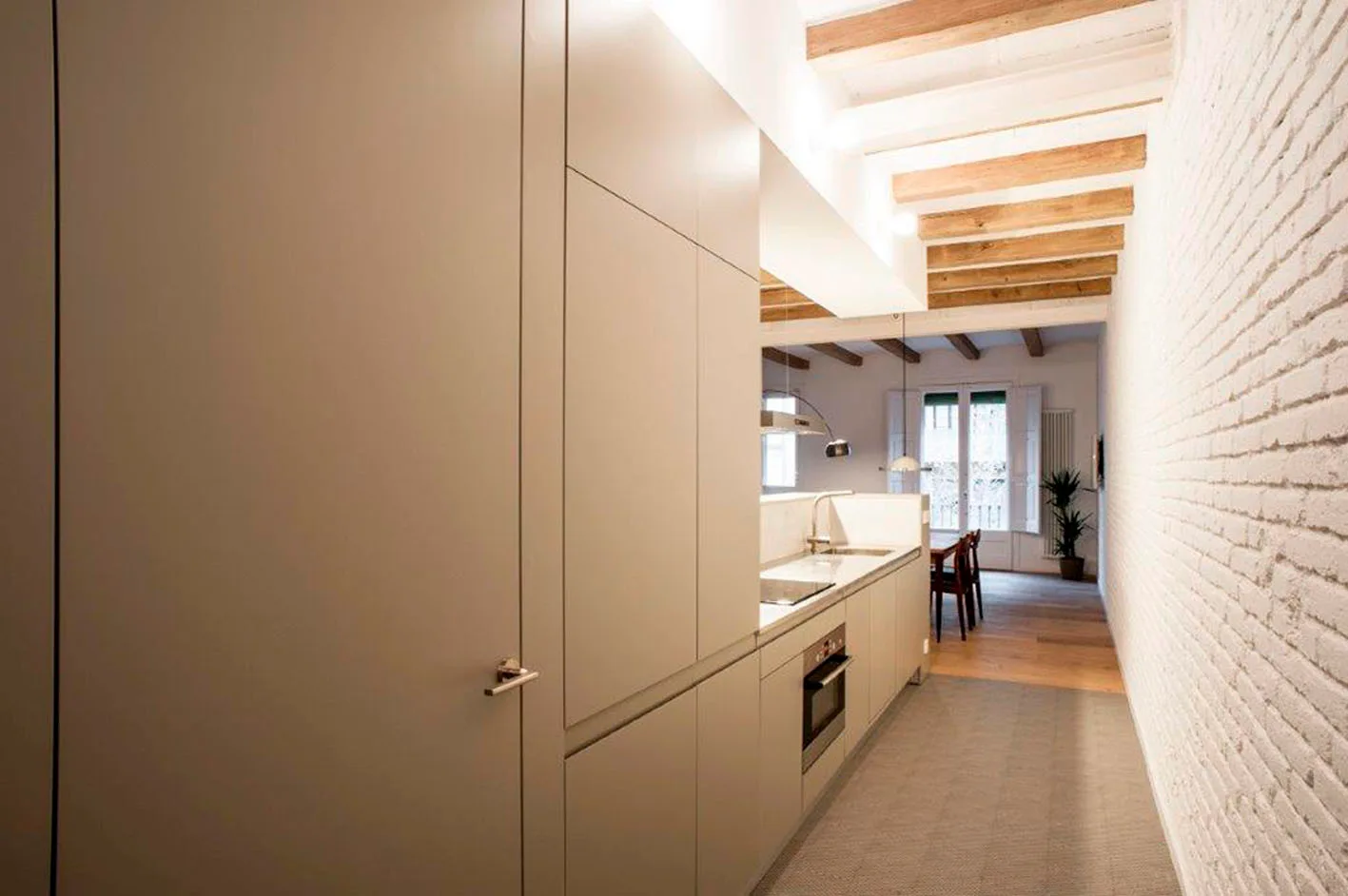 A long narrow kitchen featuring minimalist greige cabinetry, a white painted brick wall, and rustic wood ceiling beams.