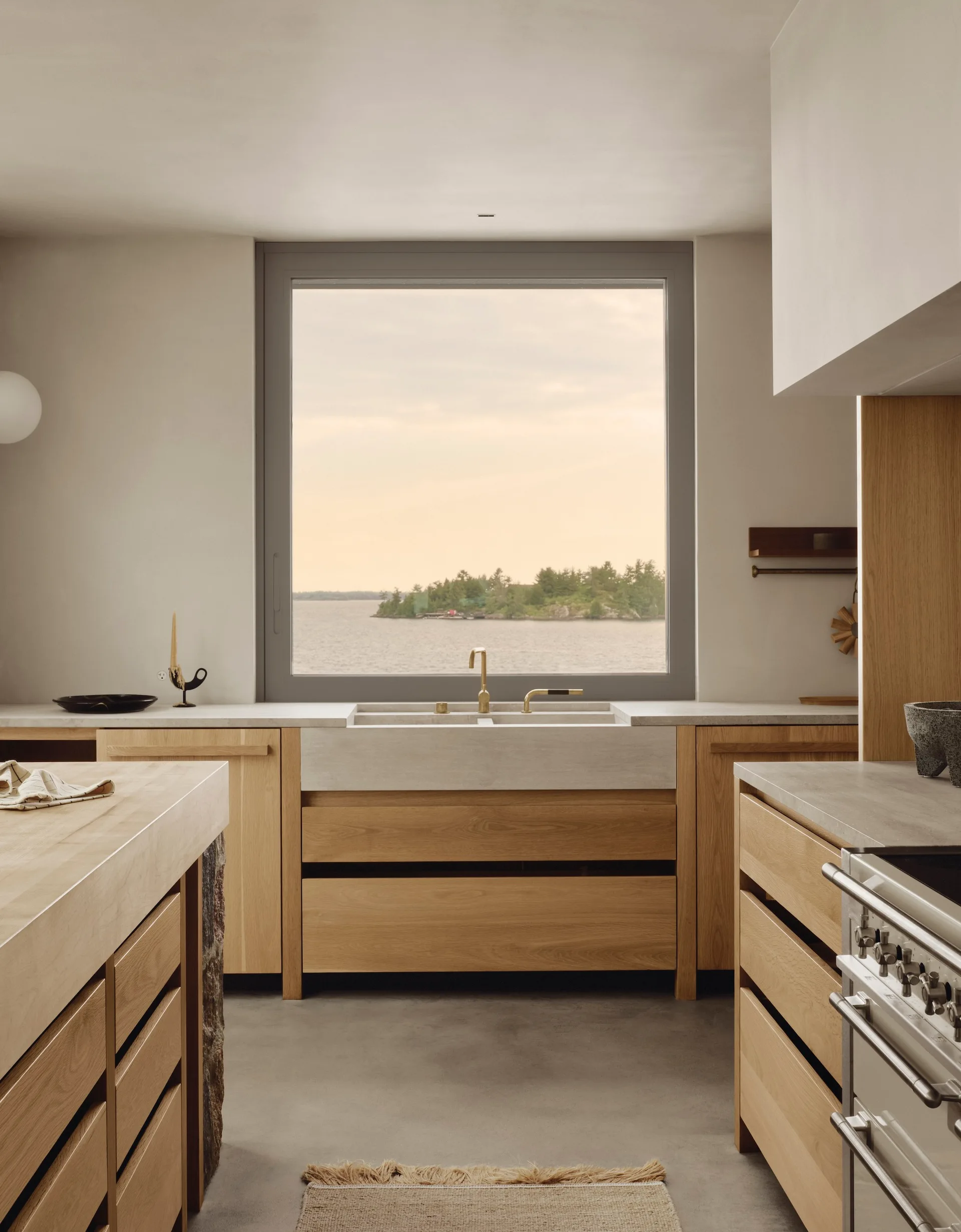 A minimalist kitchen features light oak cabinetry, a grey concrete farmhouse sink with a brass faucet, and a large window looking out onto a lake.