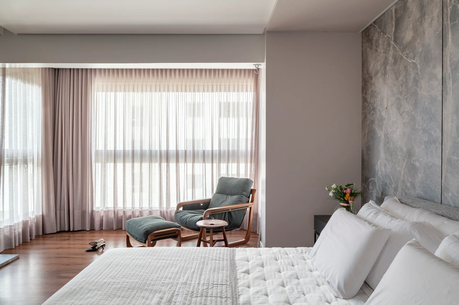 A modern bedroom features a white quilted bed against a grey veined stone feature wall and a dark green lounge chair with ottoman near a window.