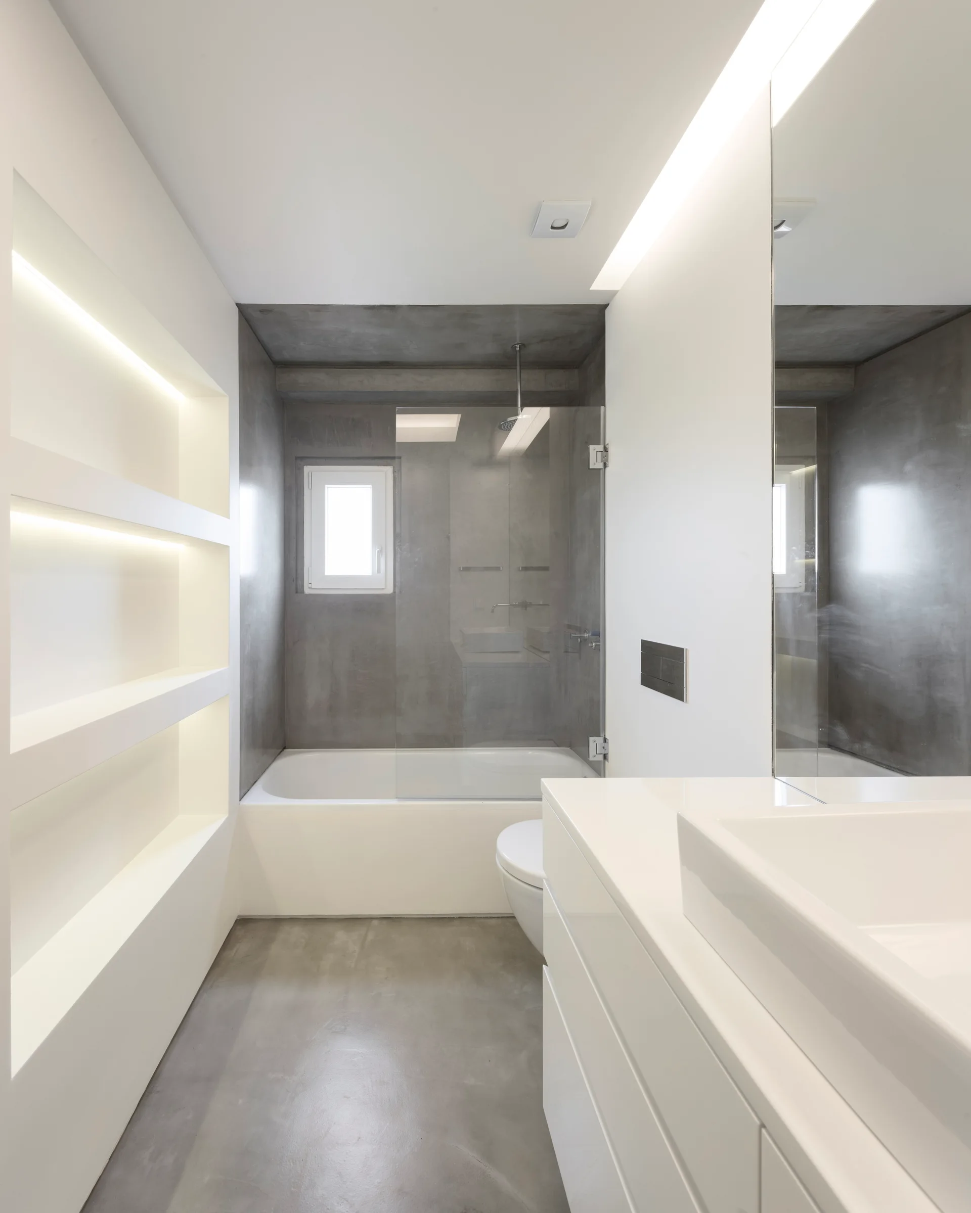 A narrow minimalist bathroom featuring a gray concrete shower surround, a white bathtub, and glowing recessed shelves on a white wall.