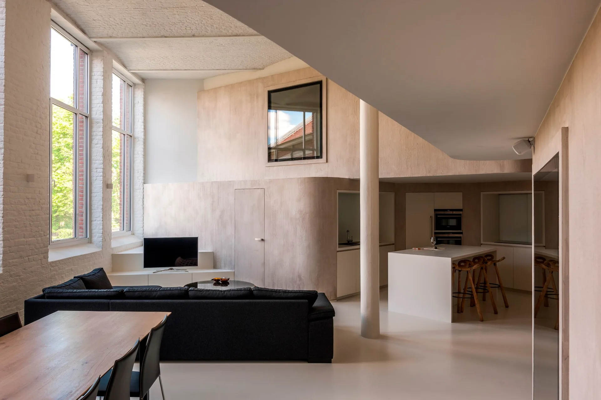 An open-concept loft features a dark grey sofa, tall industrial windows, a round structural column, and a kitchen island against curved plaster walls.